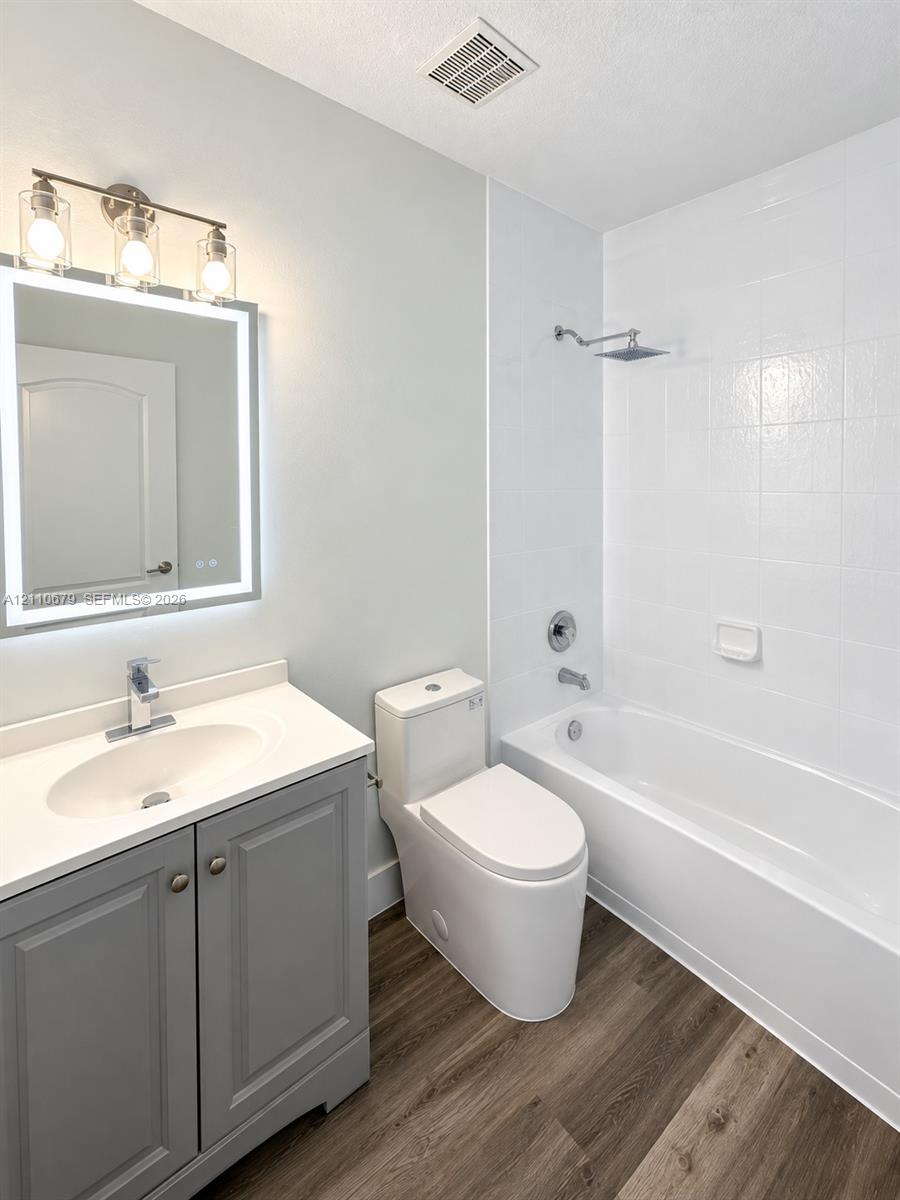 Bathroom featuring a gray vanity with a solid-surface countertop, an LED-backlit mirror, wood-finish flooring, and a bathtub with a tiled surround