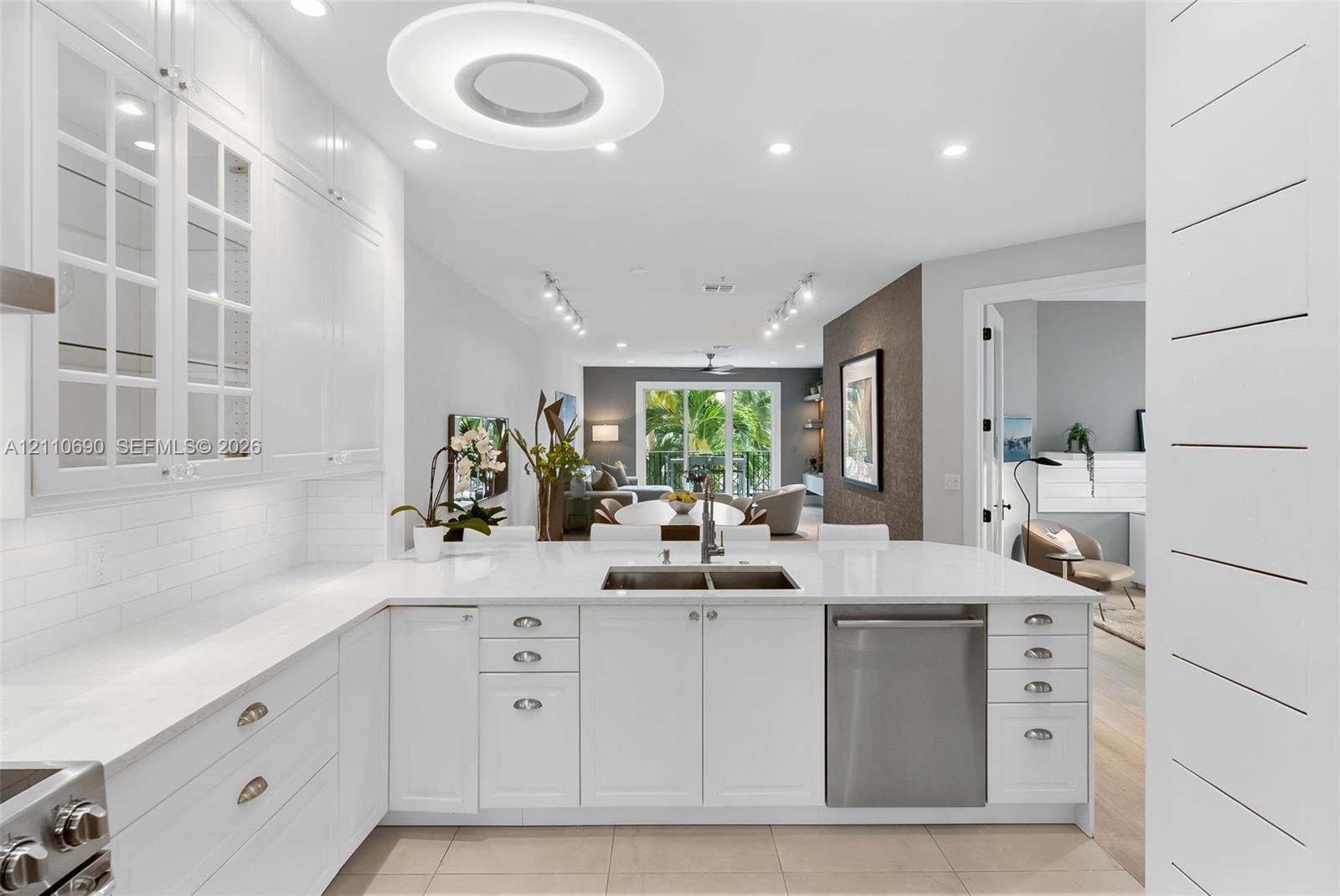 White cabinetry featuring glass-paneled upper doors, expansive countertops with an integrated stainless steel dual-basin sink, stainless steel dishwasher, tile flooring, and decorative horizontal wall paneling White cabinetry featuring glass-paneled upper doors, expansive countertops with an integrated stainless steel dual-basin sink, stainless steel dishwasher, tile flooring, and decorative horizontal wall paneling