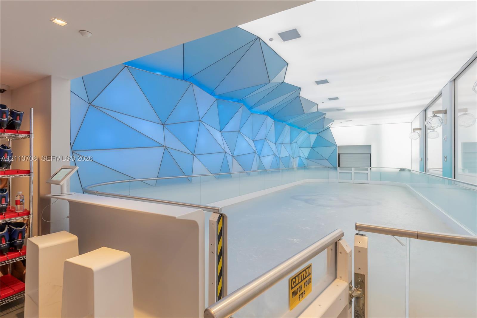 Indoor ice rink featuring a geometric wall installation with integrated blue lighting, frameless glass guardrails, and white sleek cabinetry Indoor ice rink featuring a geometric wall installation with integrated blue lighting, frameless glass guardrails, and white sleek cabinetry