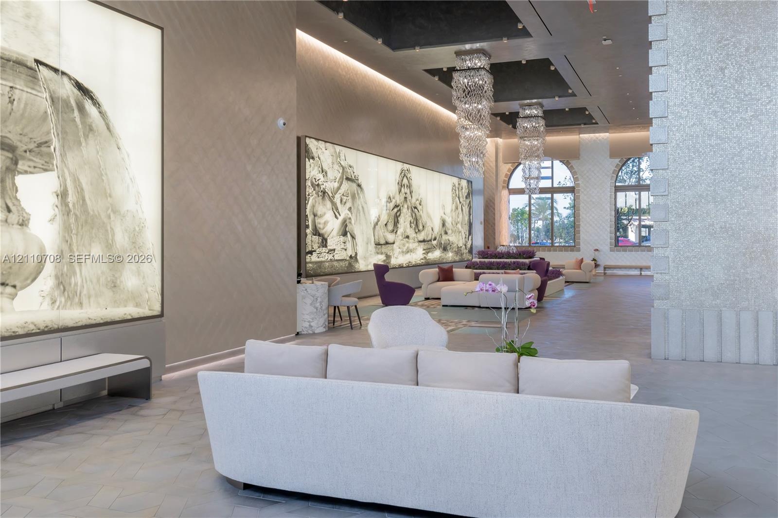 Grand interior hall featuring herringbone-pattern flooring, coffered ceiling with inset crystal chandeliers, and illuminated large-scale wall art Grand interior hall featuring herringbone-pattern flooring, coffered ceiling with inset crystal chandeliers, and illuminated large-scale wall art