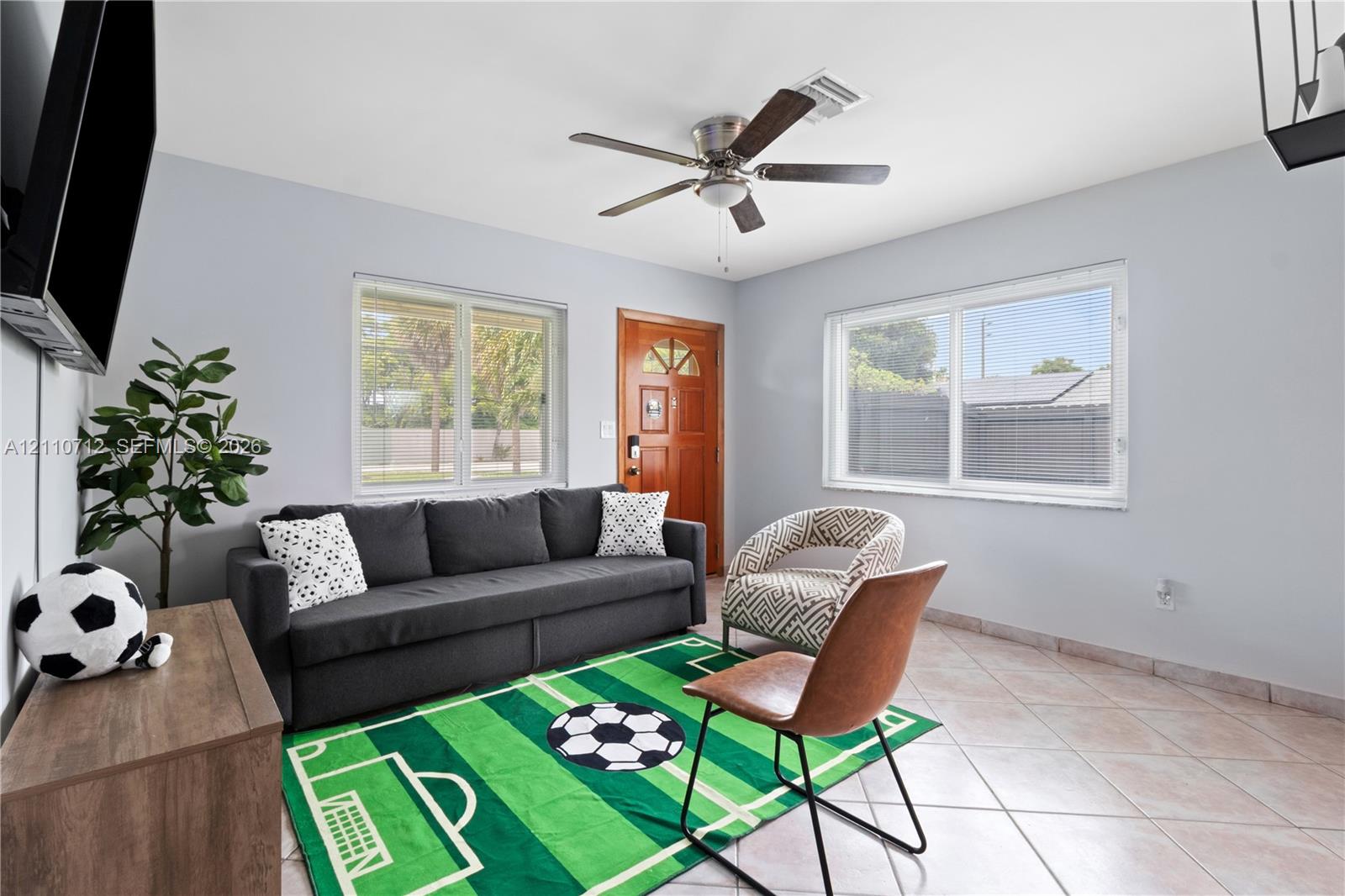 Living area featuring neutral-toned ceramic tile flooring, a multi-blade ceiling fan, and two large windows with horizontal blinds