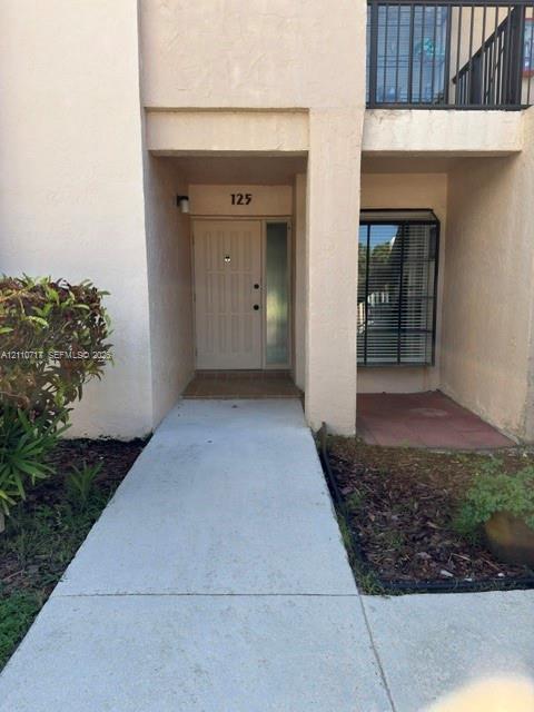 Exterior entryway featuring a concrete walkway, stucco facade, paneled door with a sidelight, and an upper-level balcony with metal railing Exterior entryway featuring a concrete walkway, stucco facade, paneled door with a sidelight, and an upper-level balcony with metal railing