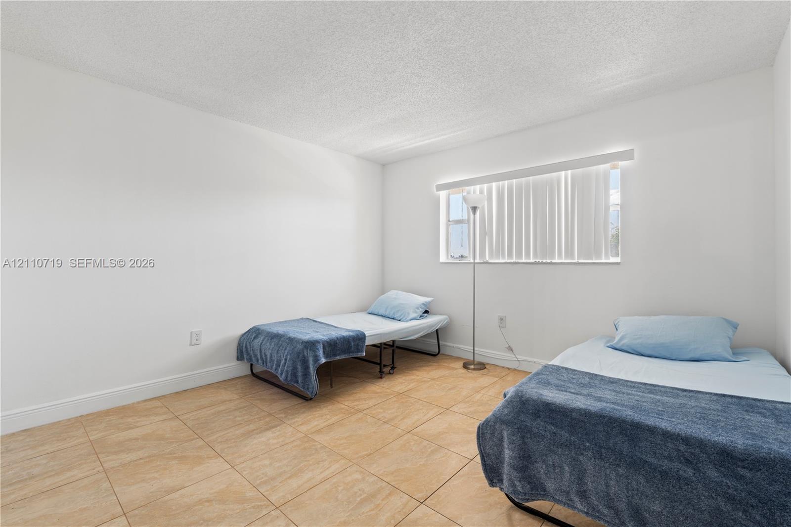 Neutral-toned tile flooring, textured ceiling, and single-pane window with horizontal blinds