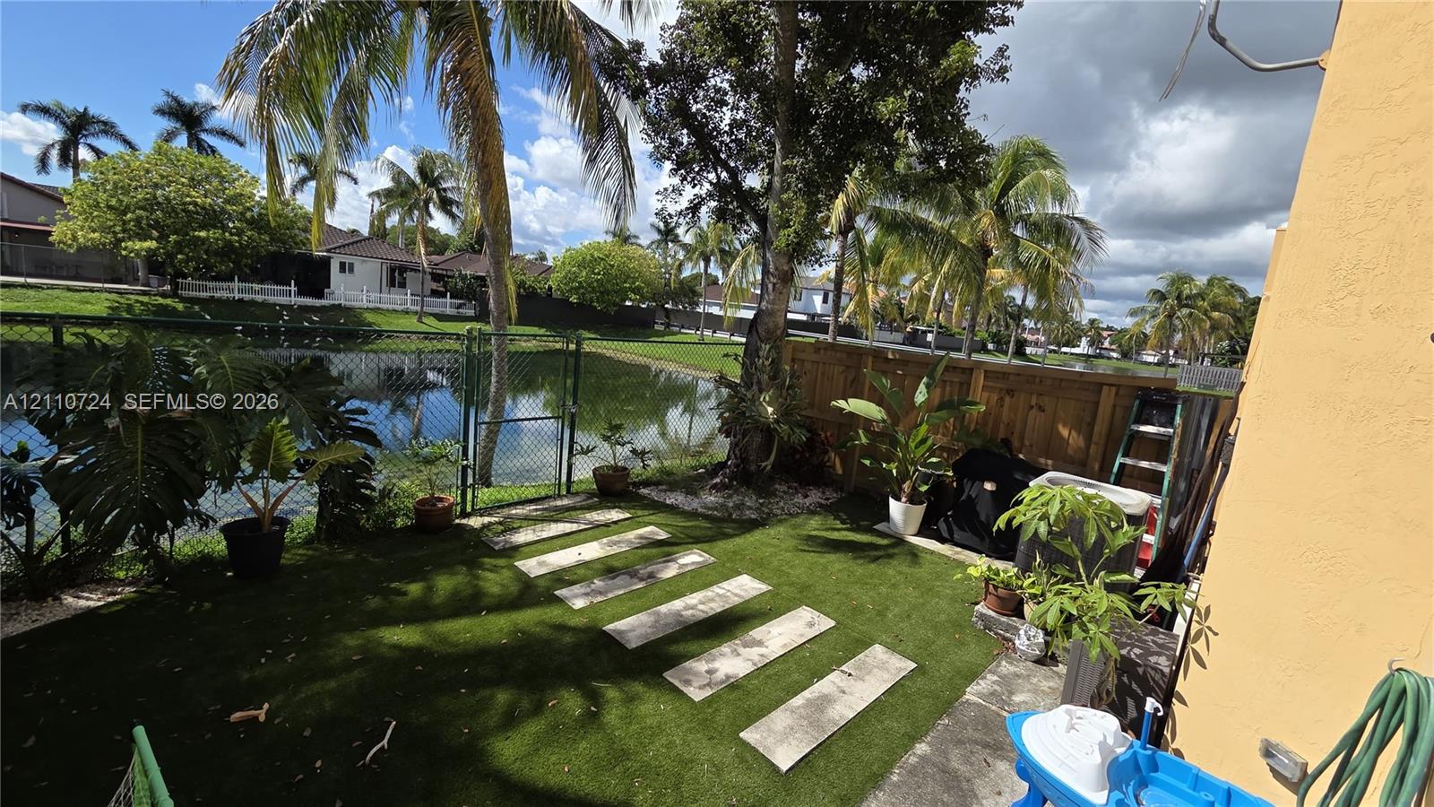 Rear patio featuring artificial turf, concrete stepping stones, a wood privacy fence, and a chain-link gate leading to the waterfront