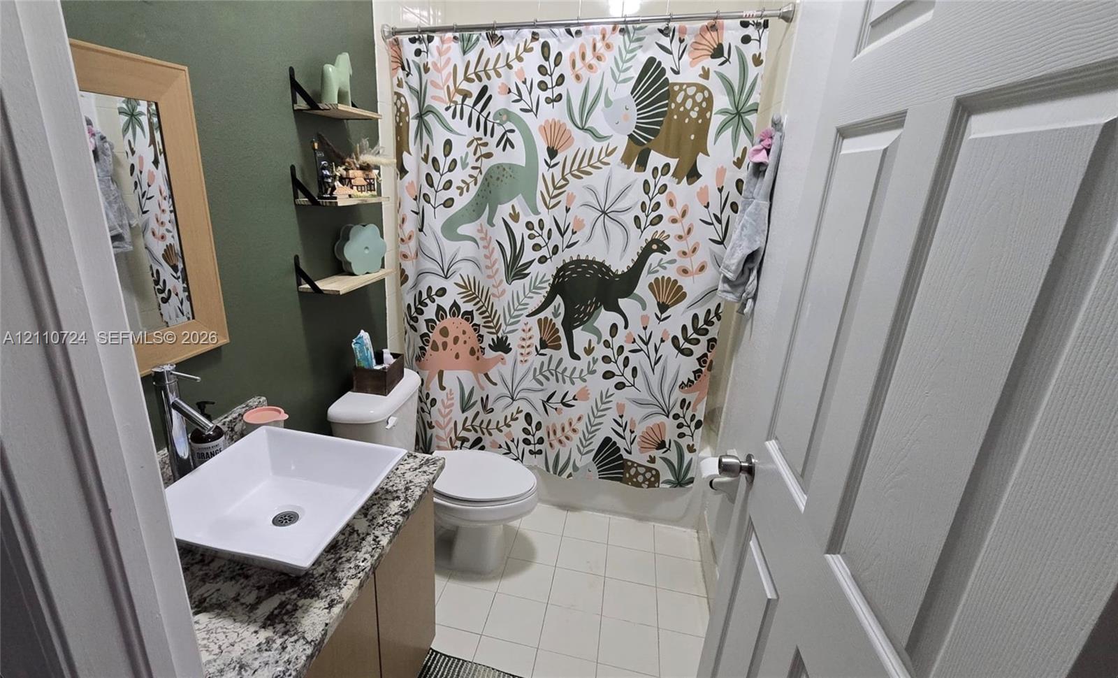 Bathroom featuring a stone-look countertop, a square vessel sink with a chrome faucet, three tiered wall-mounted corner shelves, and tile flooring