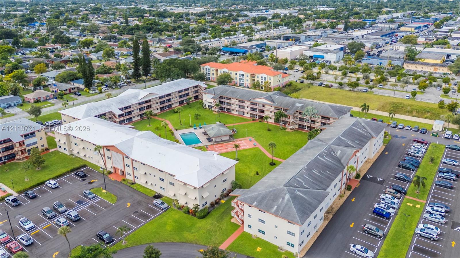 Multi-building residential complex featuring a central swimming pool, covered pavilion, paved parking areas, and mature landscaping
