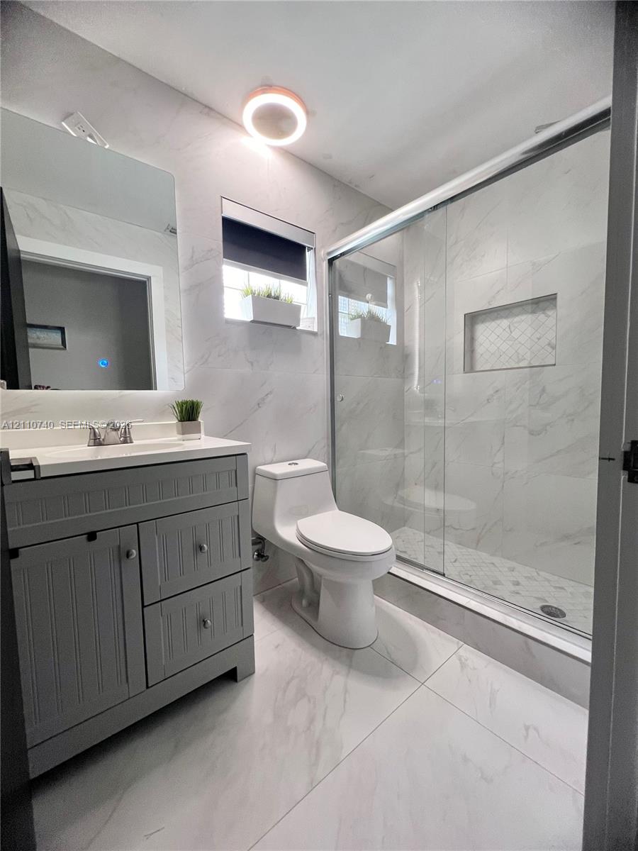 Bathroom featuring grey cabinetry with silver-tone hardware, stone-finish tiling, a glass-enclosed shower with a built-in recessed niche, and a contemporary flush-mount light fixture