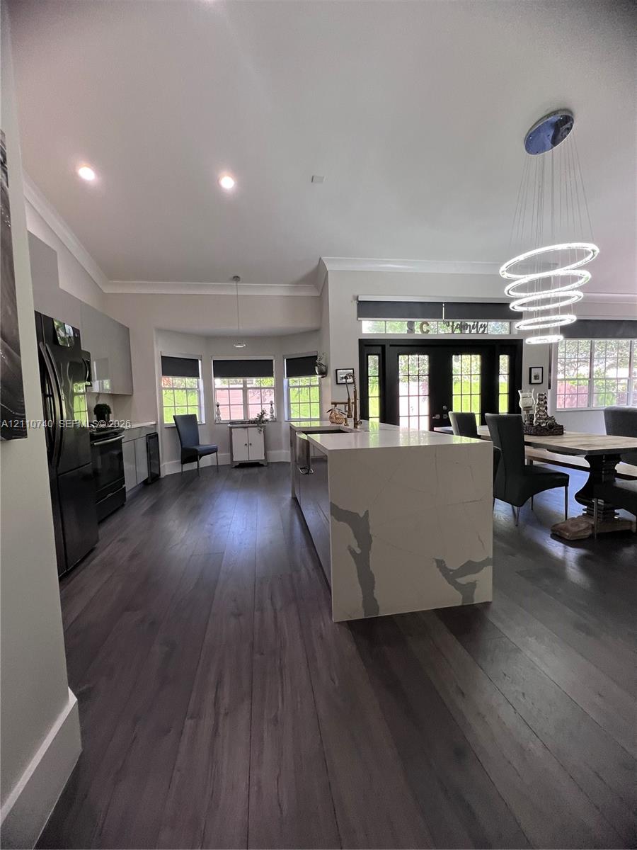 Kitchen featuring an expansive waterfall-edge island with stone-finish countertop, wood-finish flooring, recessed lighting, multi-tiered ring pendant light, and black double-entry doors with transom windows