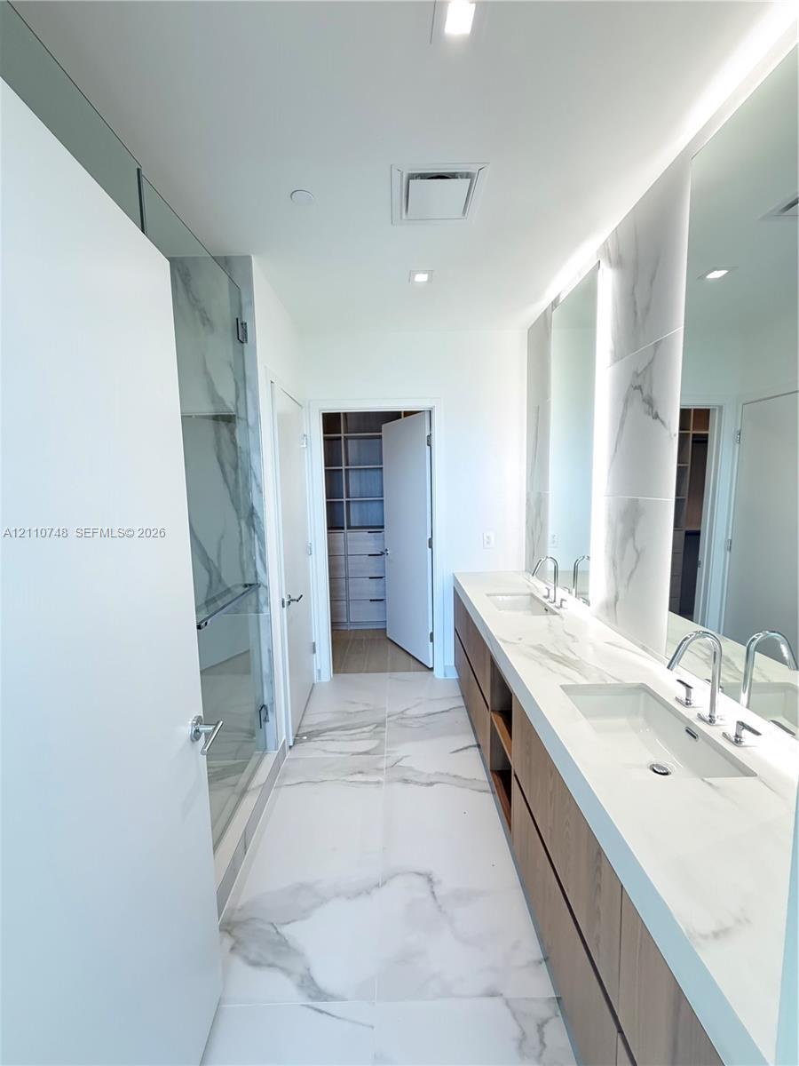 Dual-sink vanity with stone-finish countertop, full-height tile backsplash, glass-enclosed shower, and large-format porcelain tile flooring