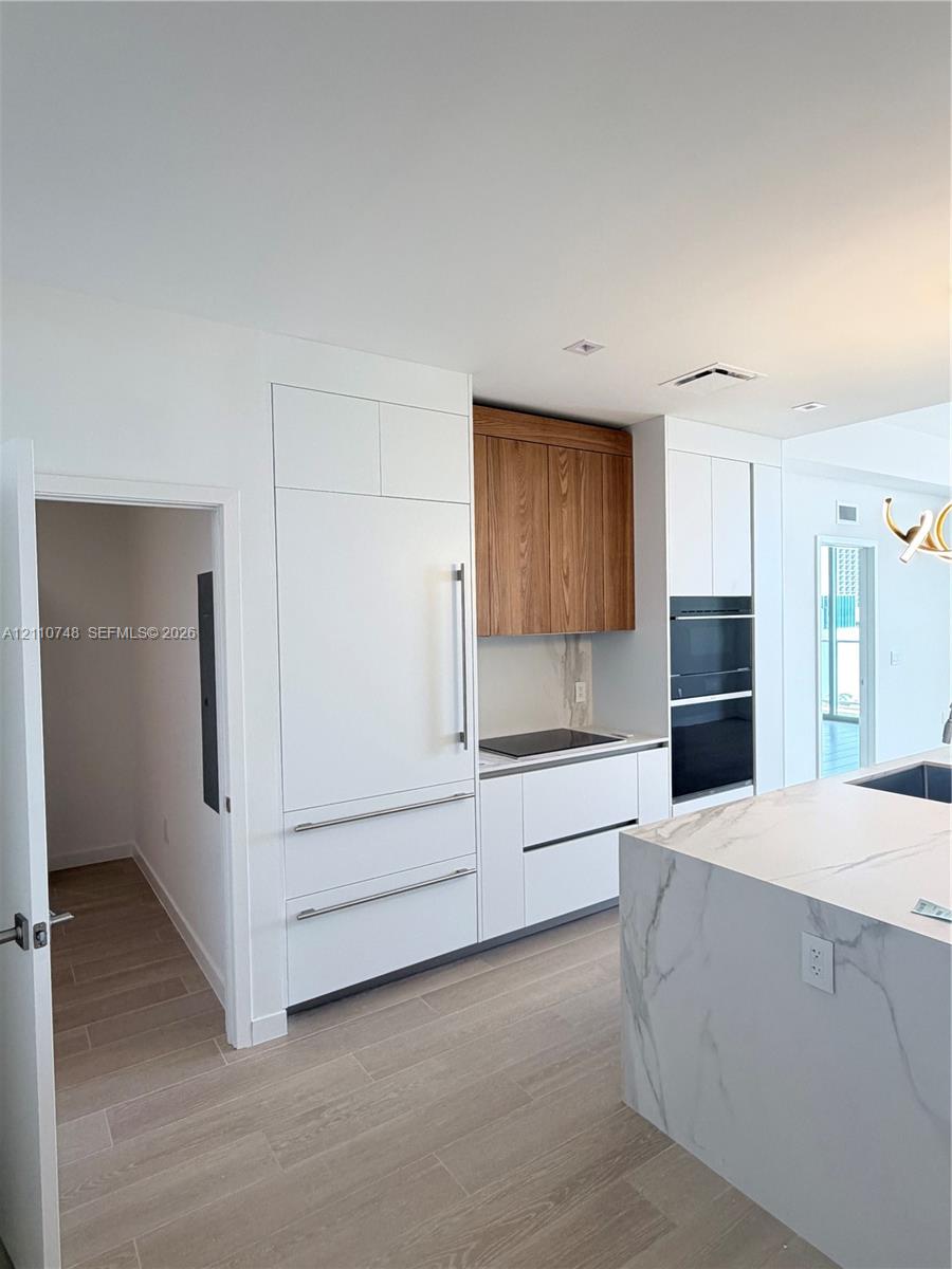 Kitchen featuring a stone-surface island, built-in integrated refrigerator, wall-mounted double oven, custom flat-panel cabinetry with wood-finish upper accents, and light-toned wood-finish flooring Kitchen featuring a stone-surface island, built-in integrated refrigerator, wall-mounted double oven, custom flat-panel cabinetry with wood-finish upper accents, and light-toned wood-finish flooring
