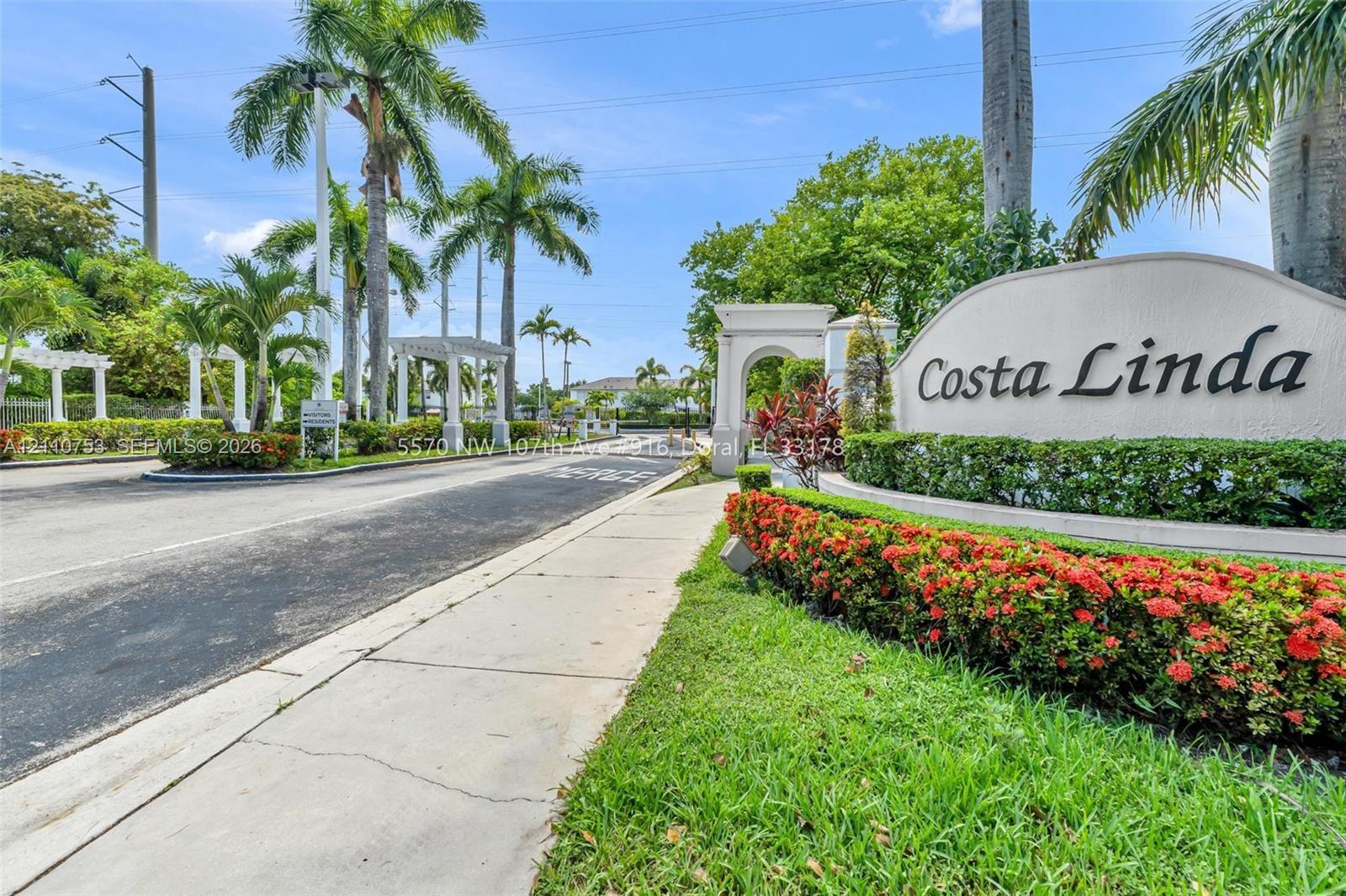 Community entrance featuring arched masonry architecture, paved roadways, mature palm trees, and manicured shrubbery