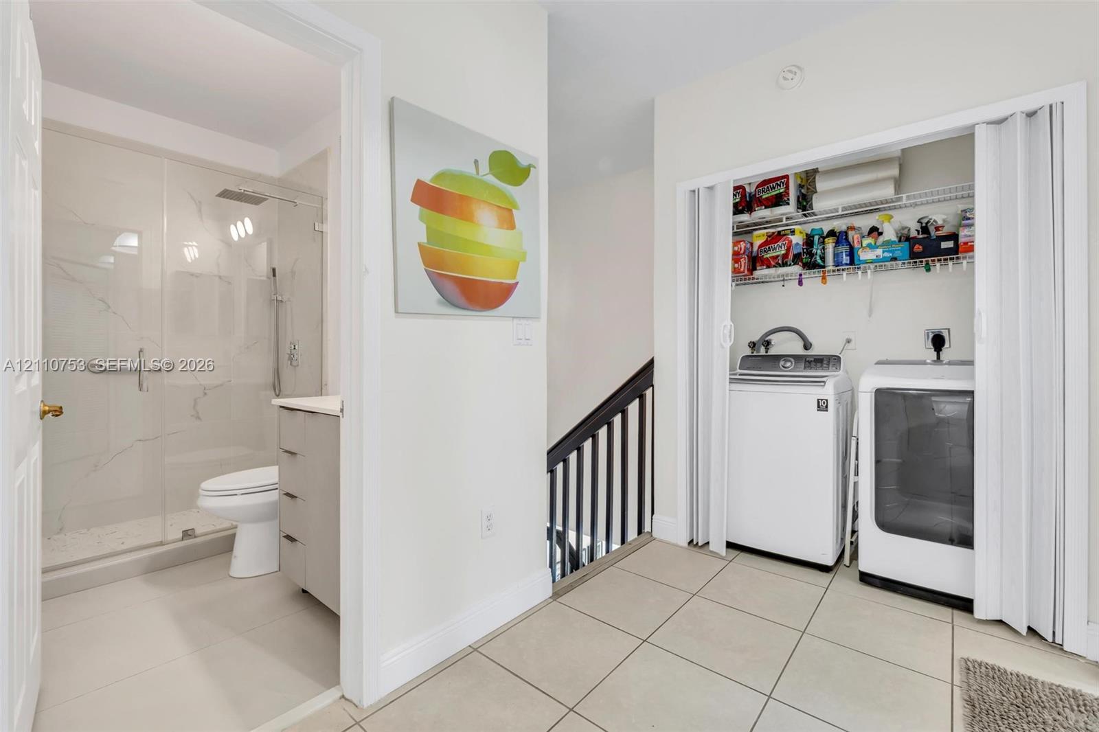 Bathroom featuring a glass-enclosed shower with a rainfall showerhead and stone-finish wall tile, complemented by a laundry closet with bi-fold doors and overhead wire shelving