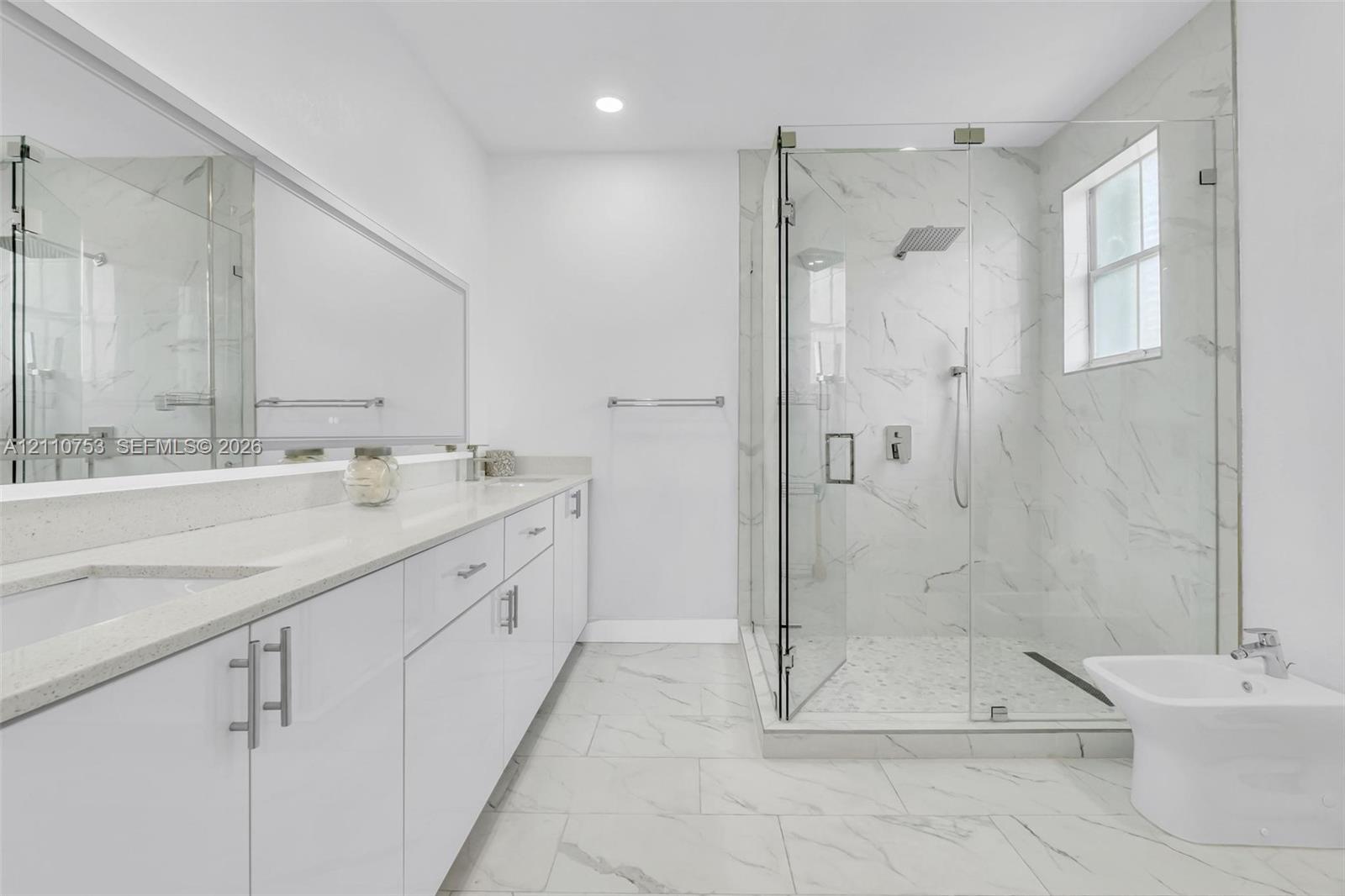 Bathroom featuring a double-sink vanity with light-toned solid surface countertops, floor-to-ceiling tiled glass-enclosed shower, large wall-mounted mirror, and tile flooring