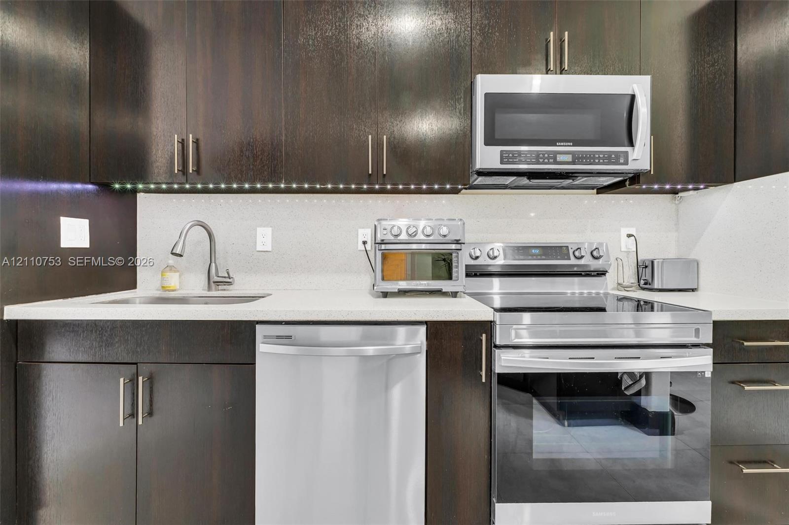 Kitchen featuring dark wood-finish cabinetry, solid surface countertops, stainless steel appliances, under-cabinet lighting, and a gooseneck faucet