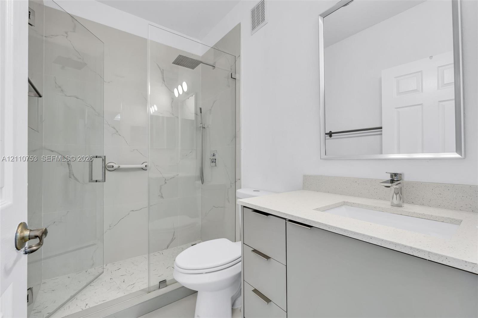 Modern bathroom featuring a light gray vanity with a stone-composite countertop, undermount sink, walk-in shower with floor-to-ceiling large-format tile, frameless glass enclosure, and a silver-framed mirror