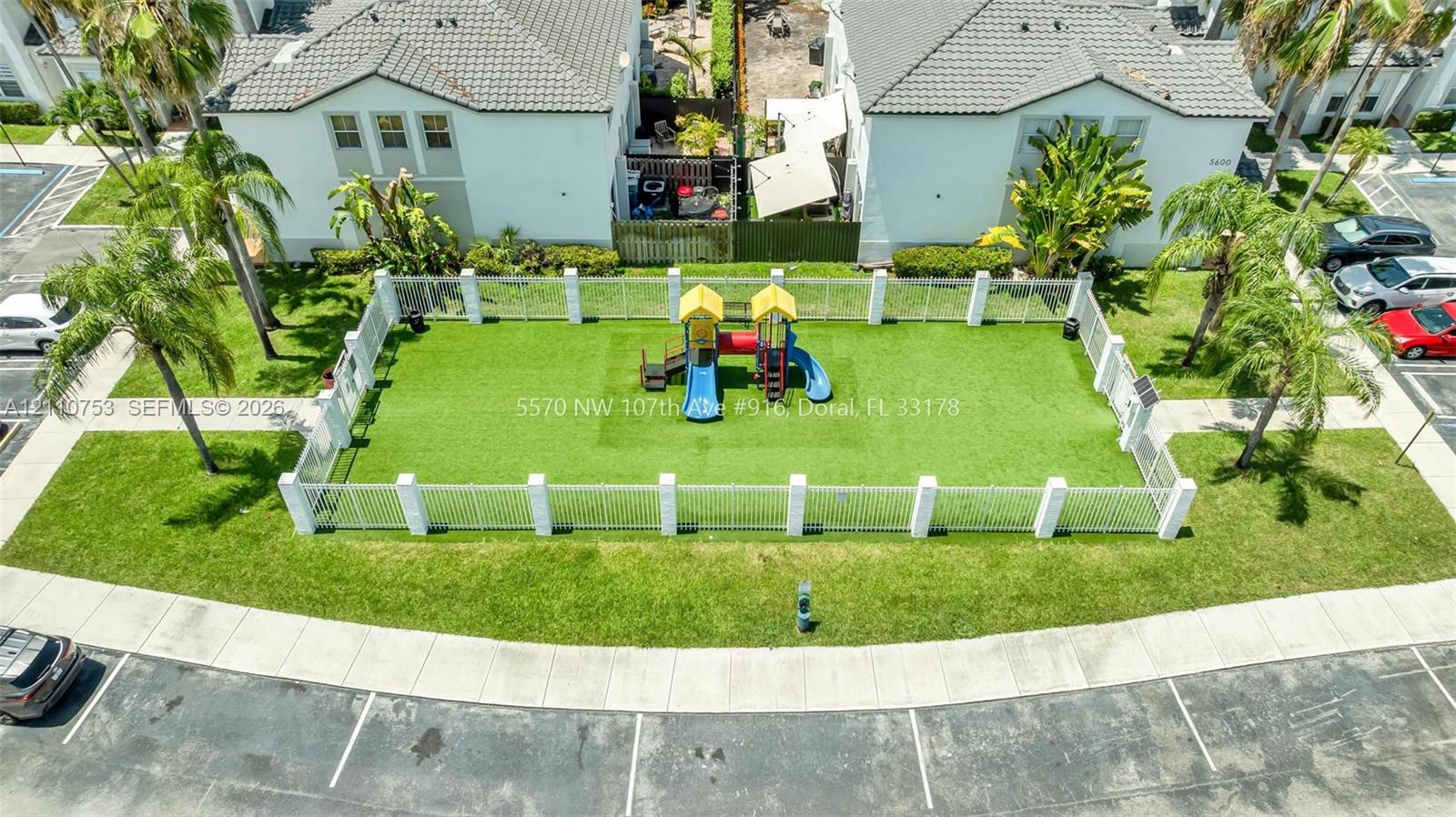 Fenced recreational area featuring a synthetic turf surface, playground structure, concrete perimeter wall, and mature palm trees