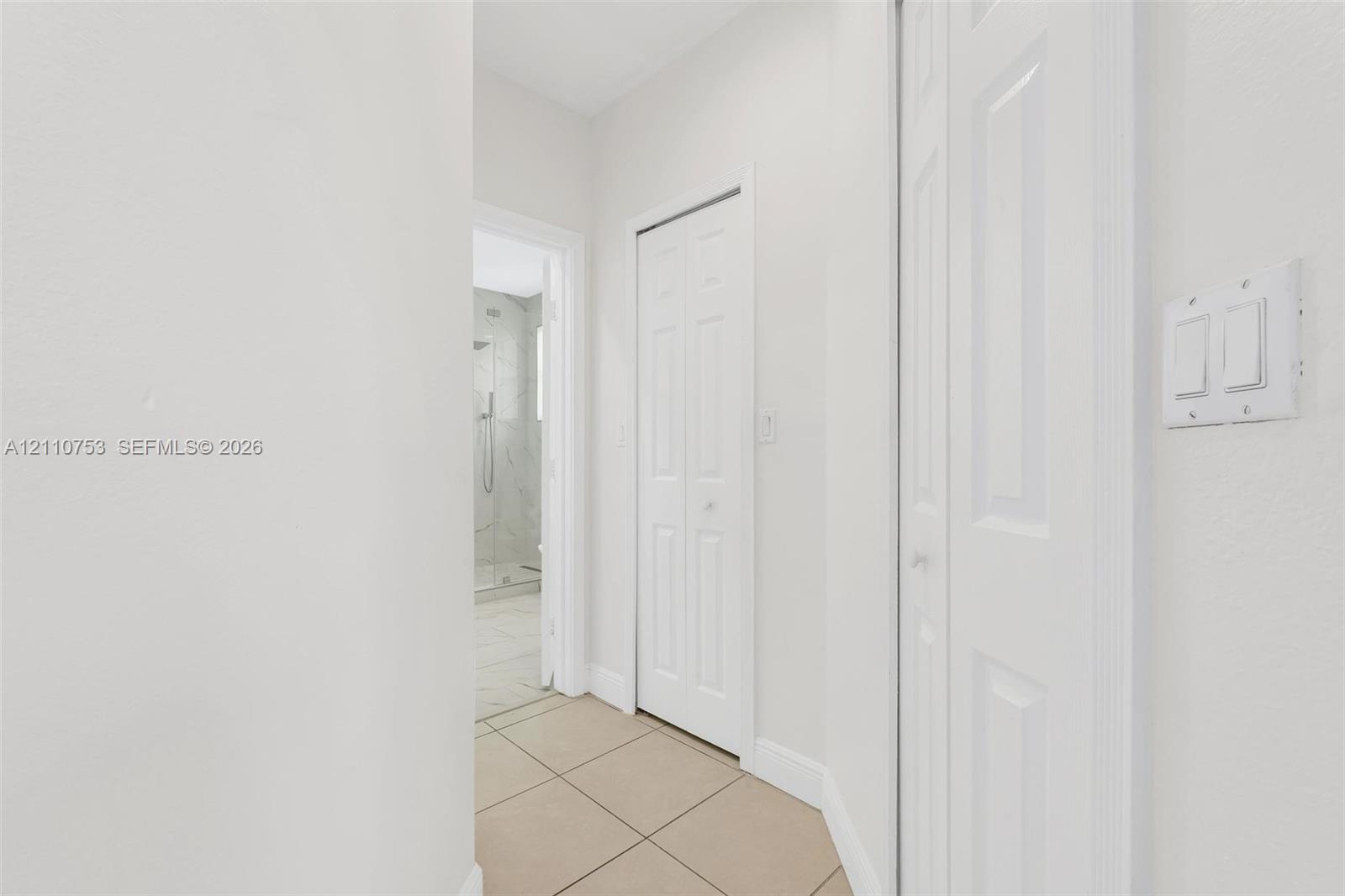 Hallway featuring neutral-toned tile flooring, two bi-fold doors, and a doorway revealing a tiled shower enclosure