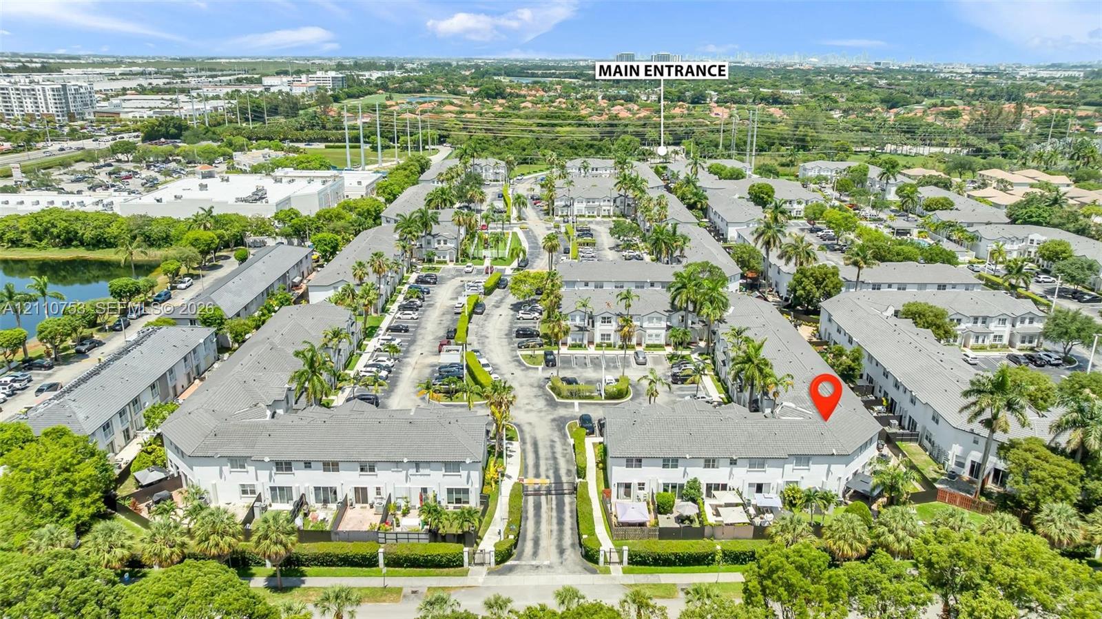 Aerial perspective of a multi-unit residential community featuring pitched shingle roofing, paved roadways, mature palm tree landscaping, and perimeter hedge screening