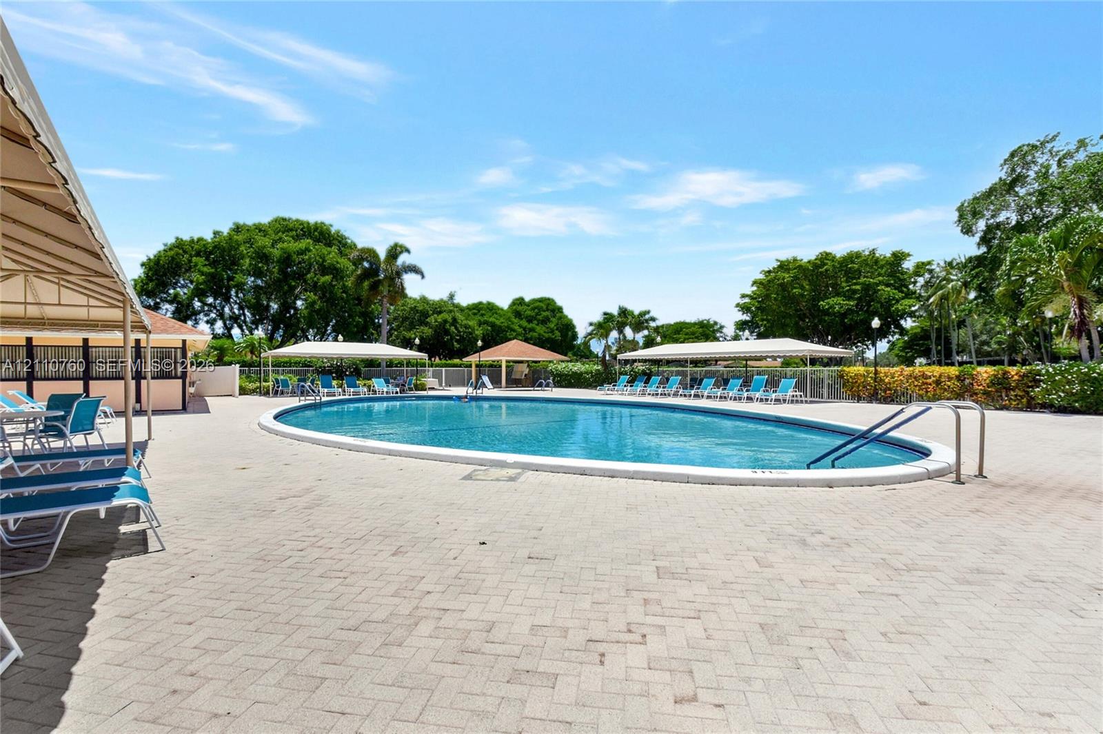 Oval swimming pool with a paved deck, multiple shade structures, and mature perimeter landscaping