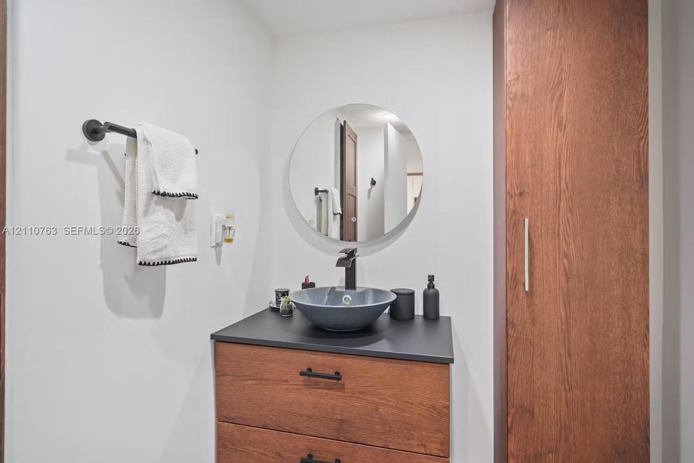Modern vanity featuring a wood-finish cabinet, dark solid-surface countertop, circular vessel sink, and matte black faucet, complemented by a tall built-in storage cabinet and a circular wall-mounted mirror Modern vanity featuring a wood-finish cabinet, dark solid-surface countertop, circular vessel sink, and matte black faucet, complemented by a tall built-in storage cabinet and a circular wall-mounted mirror