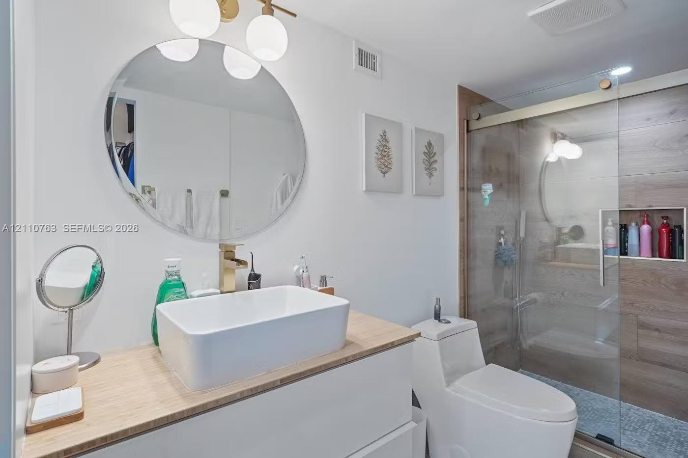 Floating vanity with a light wood-finish countertop, rectangular vessel sink, gold-tone faucet, round wall-mounted mirror, and glass-enclosed shower with wood-finish tile wall and built-in recessed niche Floating vanity with a light wood-finish countertop, rectangular vessel sink, gold-tone faucet, round wall-mounted mirror, and glass-enclosed shower with wood-finish tile wall and built-in recessed niche