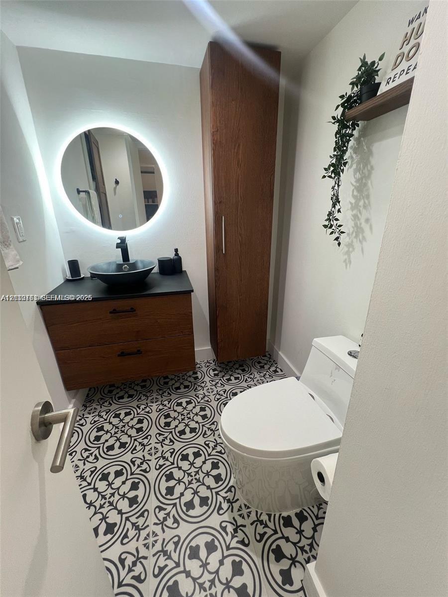 Modern vanity with wood-finish cabinetry, black stone countertop, and circular vessel sink, complemented by a backlit round mirror, floor-to-ceiling wood-finish storage cabinet, and patterned tile flooring Modern vanity with wood-finish cabinetry, black stone countertop, and circular vessel sink, complemented by a backlit round mirror, floor-to-ceiling wood-finish storage cabinet, and patterned tile flooring