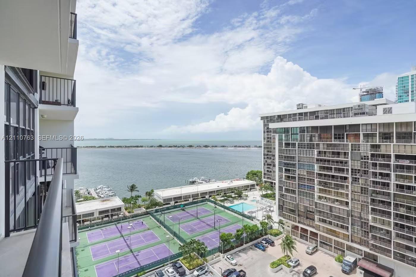 Covered balcony with black metal railings, overlooking tennis courts, a swimming pool, and a coastal waterway