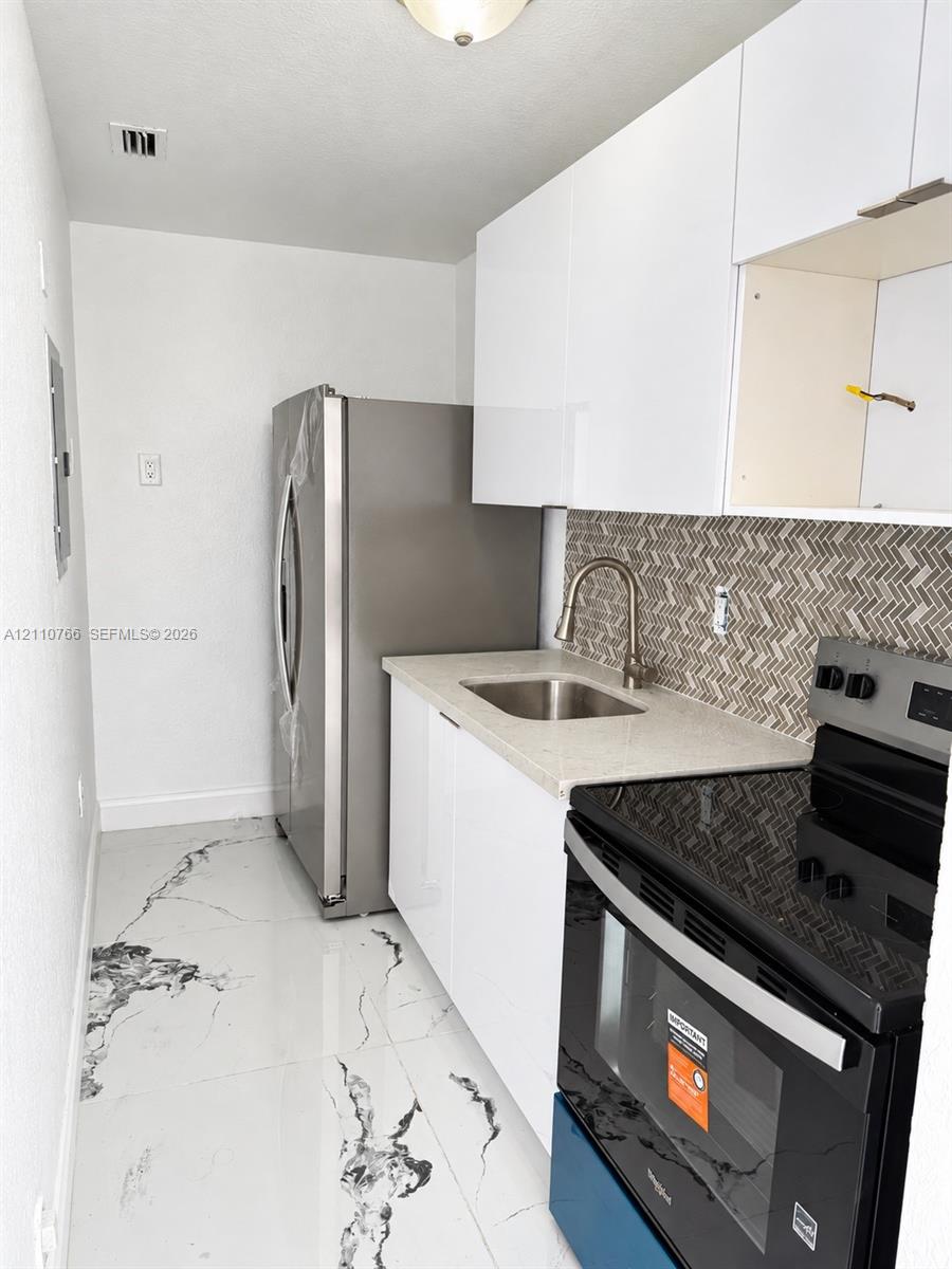 Kitchen featuring white gloss cabinetry, stone-finish countertops, stainless steel refrigerator, black finish range, and herringbone tile backsplash over marble-finish flooring Kitchen featuring white gloss cabinetry, stone-finish countertops, stainless steel refrigerator, black finish range, and herringbone tile backsplash over marble-finish flooring