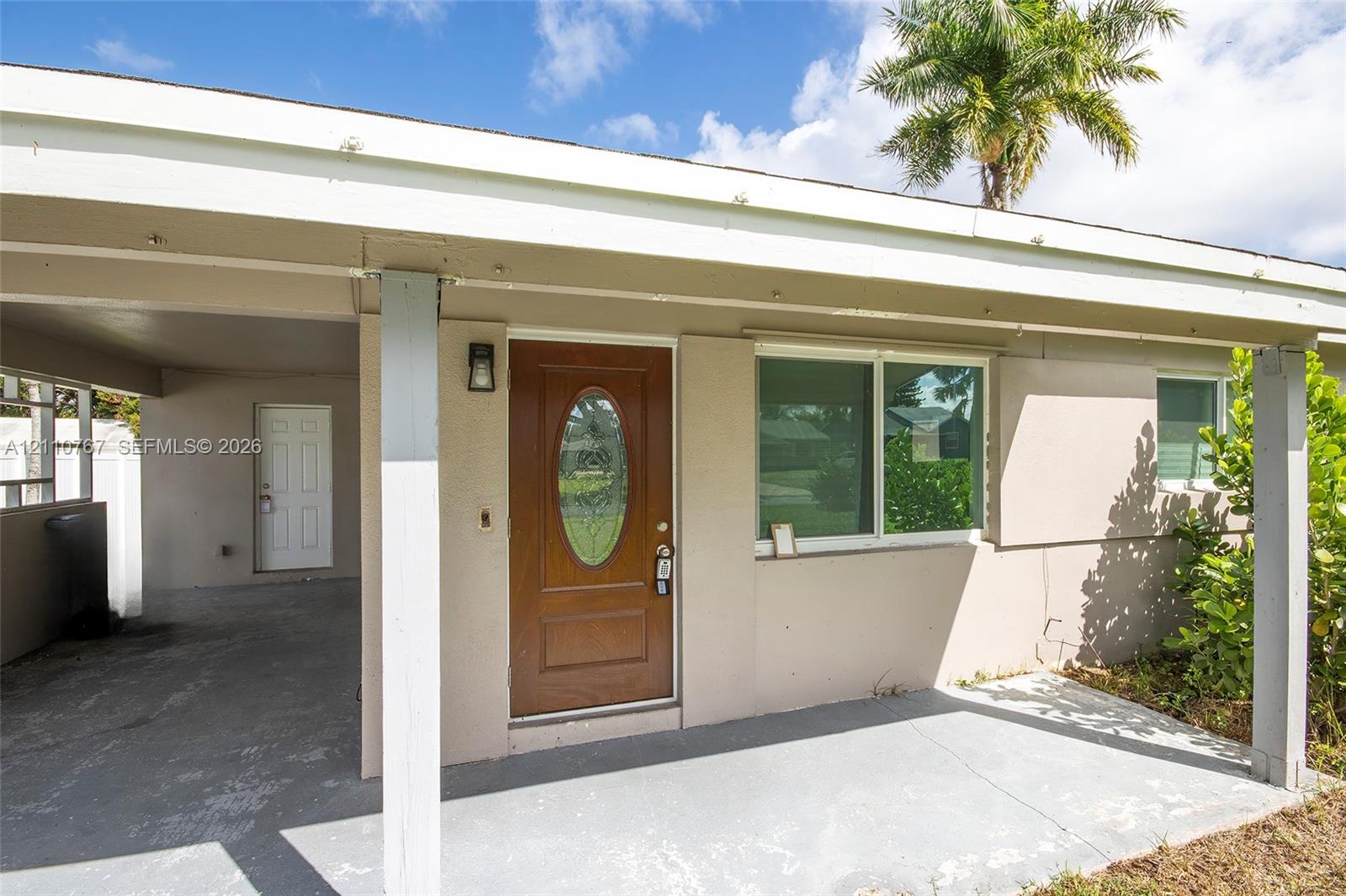 Exterior entryway featuring a covered carport, wood-finished front door with an oval glass inlay, sliding window with horizontal muntins, and a concrete porch