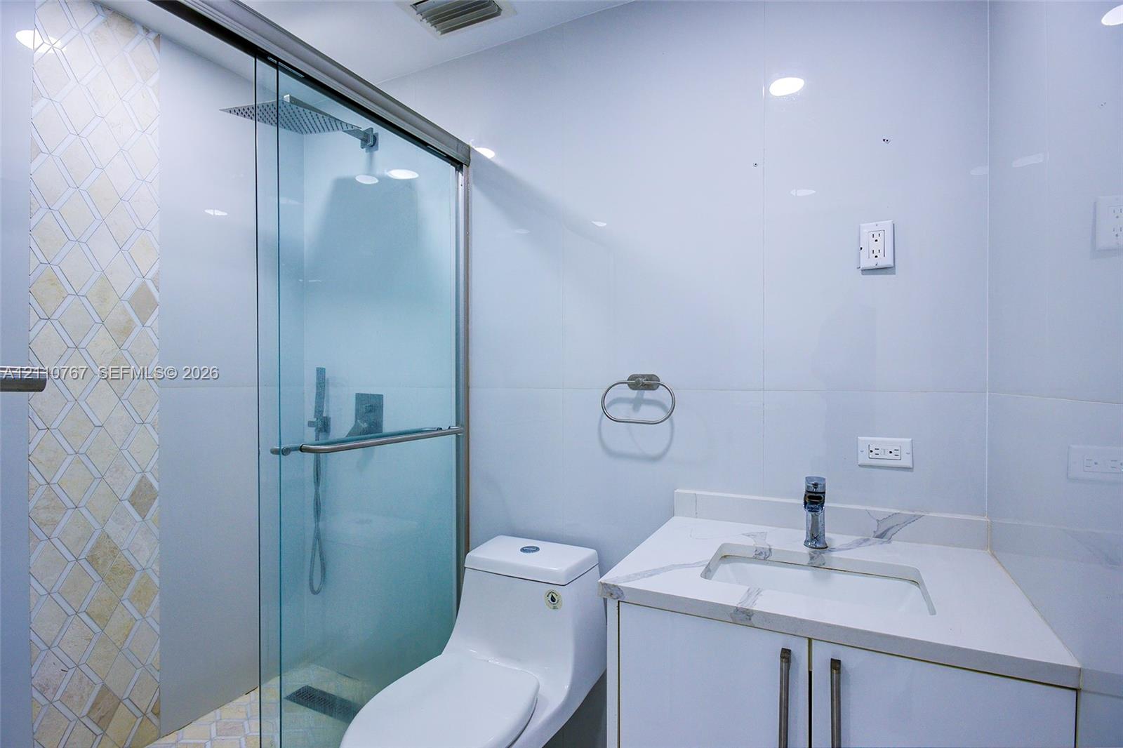 Walk-in glass shower enclosure with a rainfall showerhead, white vanity cabinet with an integrated rectangular sink, wall-mounted faucet, and decorative tiled shower wall accent