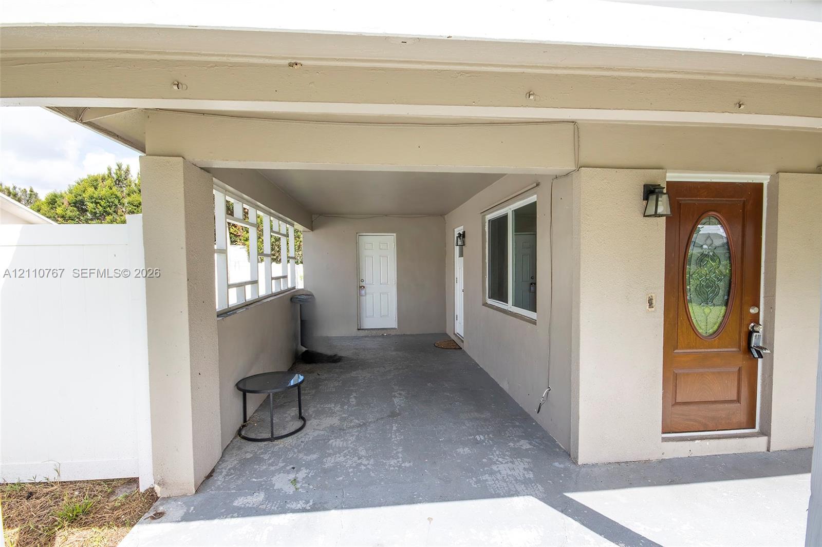 Covered carport with concrete flooring, solid wood-finish entry door featuring a glass oval inlay, and exterior wall-mounted lighting