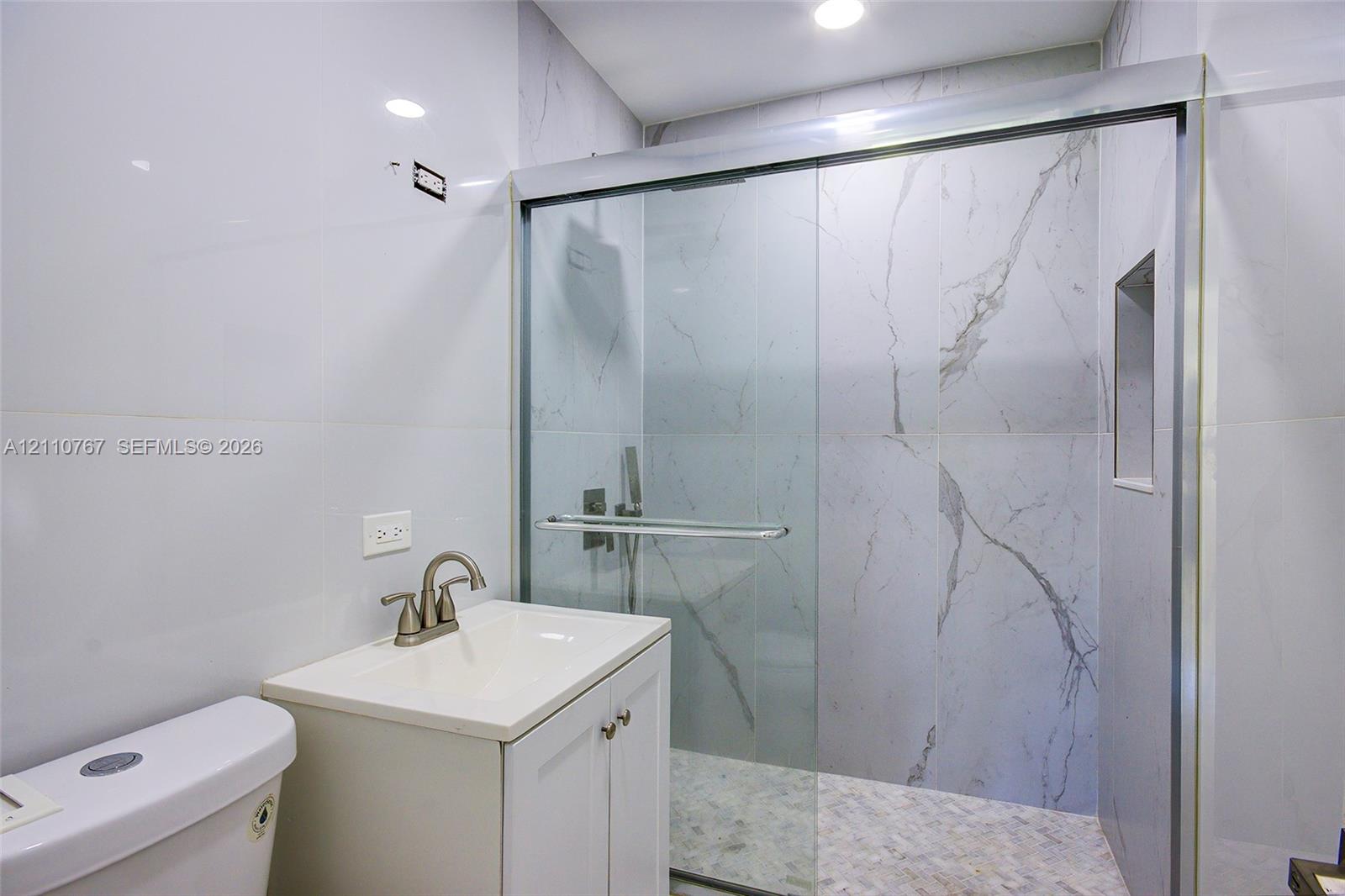 Full-height stone-finish wall tile, frameless glass shower enclosure with sliding door, integrated shower niche, modern vanity cabinet with drop-in sink, and brushed-finish faucet
