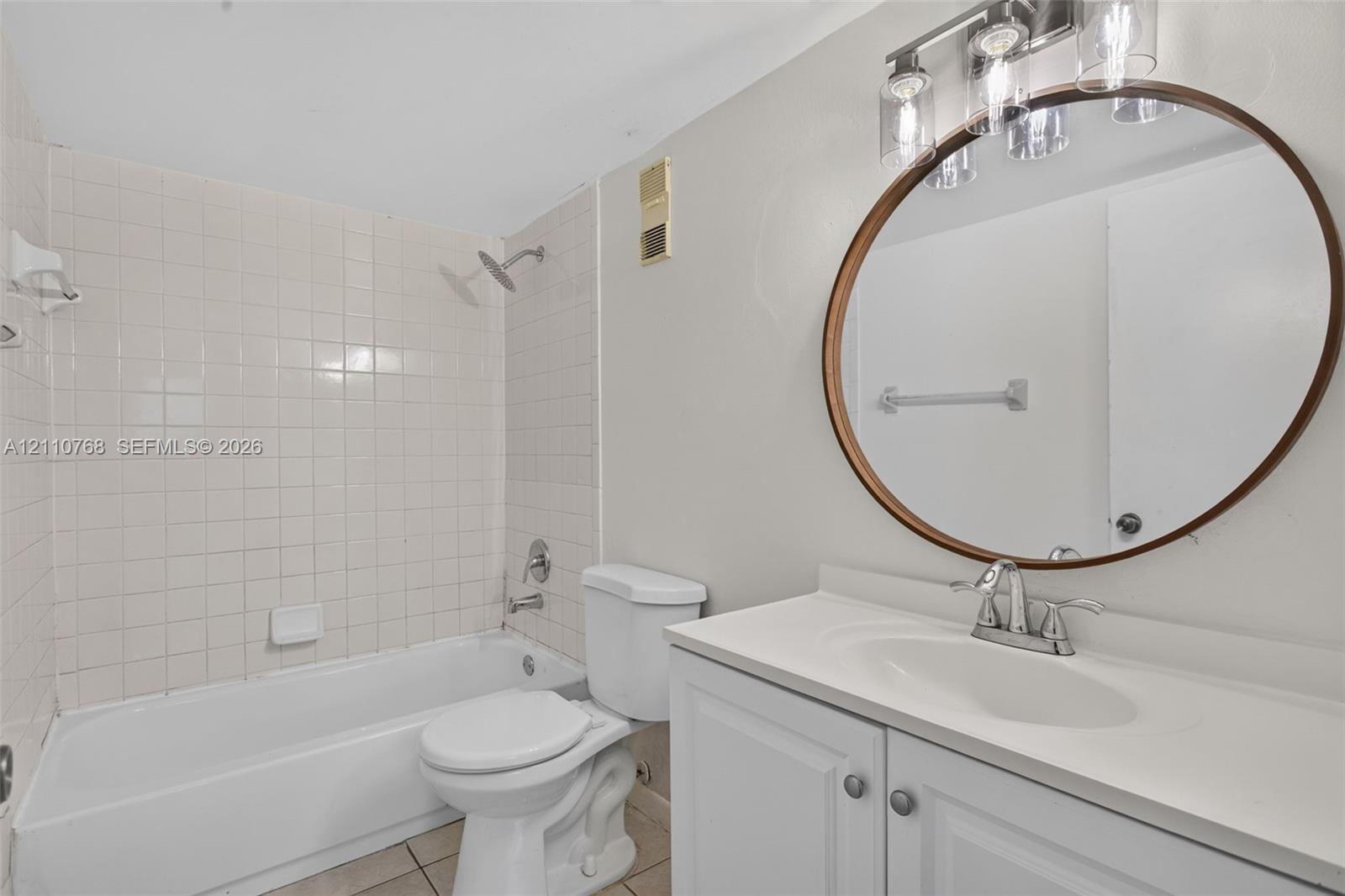 Bathroom featuring a white vanity cabinet with a solid-surface countertop, circular wall-mounted mirror, three-light fixture, and a tiled shower-tub combination