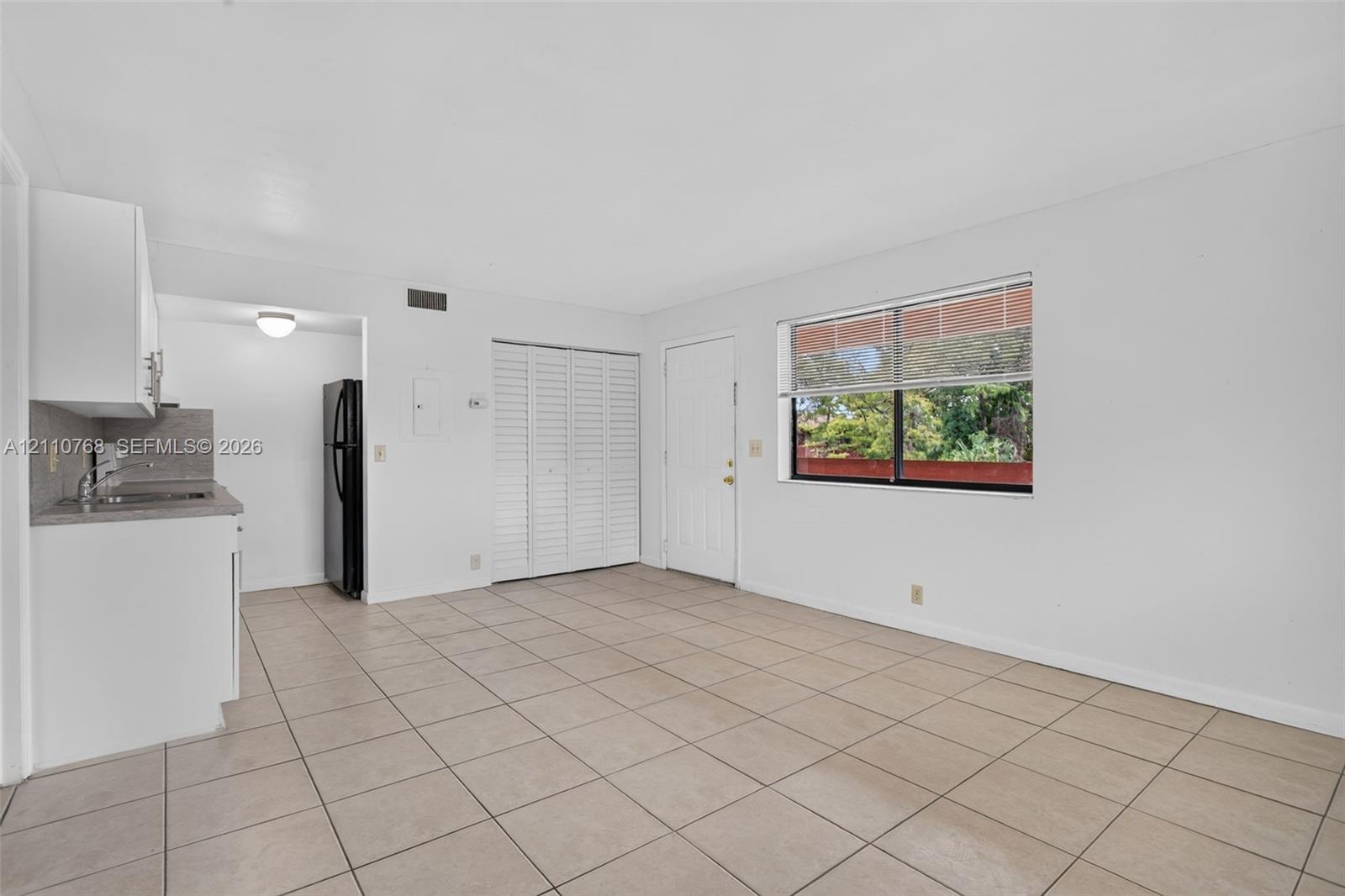 Open living area featuring neutral tile flooring, white cabinetry, louvered closet doors, and large windows with integrated blinds