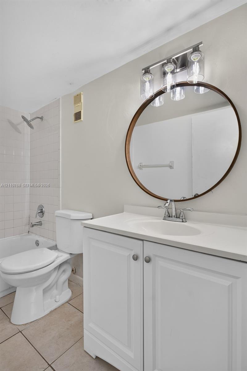 White vanity with a solid-surface countertop and integrated sink, round mirror with a bronze-toned frame, three-light fixture with glass shades, and tiled tub surround