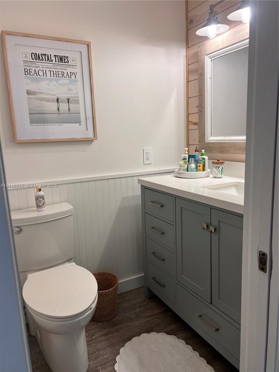 Bathroom featuring a vanity with soft-toned cabinetry, light-colored solid surface countertop, white beadboard wainscoting, wood-finish flooring, and horizontal wood-plank accent wall
