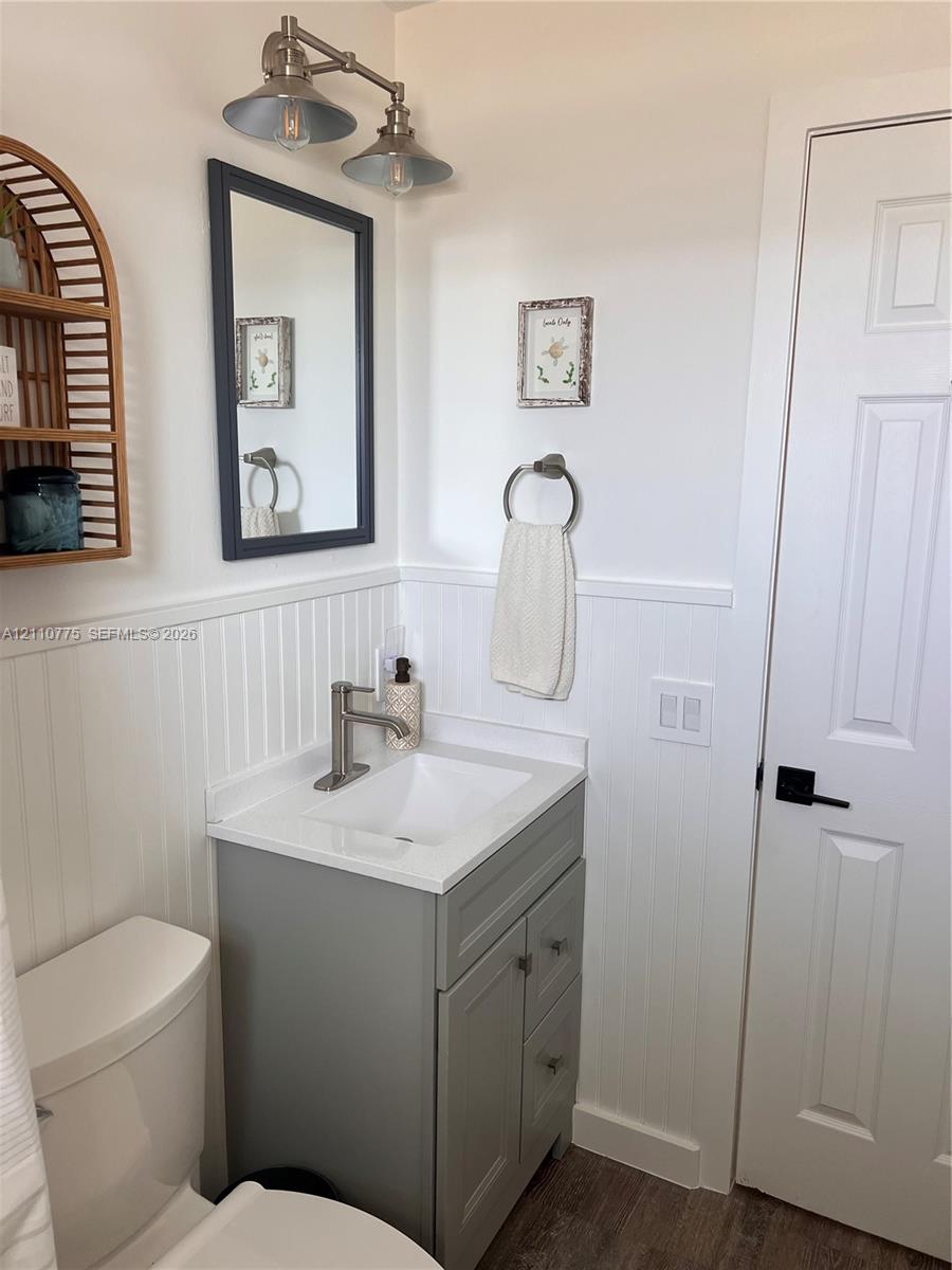 Gray vanity with a white countertop and basin, white beadboard wainscoting, dual-light metal vanity fixture, framed wall mirror, and six-panel interior door with matte black hardware