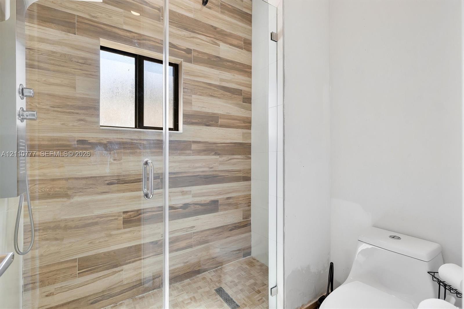 Walk-in shower featuring wood-finish tile wall cladding, frameless glass enclosure, wall-mounted shower fixture, and a built-in window, paired with a one-piece toilet