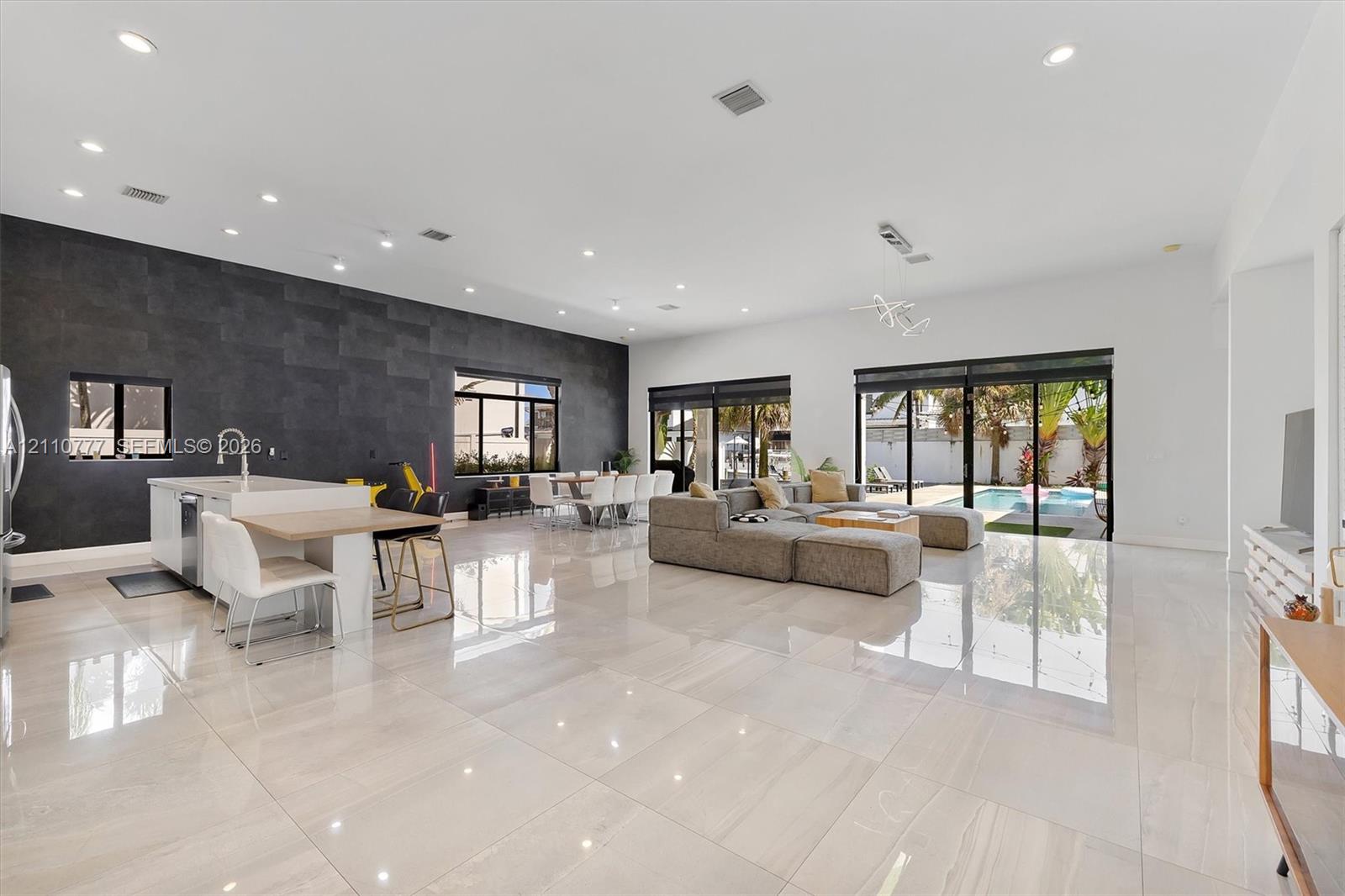 Open-concept living space featuring high-gloss tile flooring, recessed lighting, a statement dark tile accent wall, an island kitchen with built-in cabinetry, and multi-panel glass sliding doors Open-concept living space featuring high-gloss tile flooring, recessed lighting, a statement dark tile accent wall, an island kitchen with built-in cabinetry, and multi-panel glass sliding doors