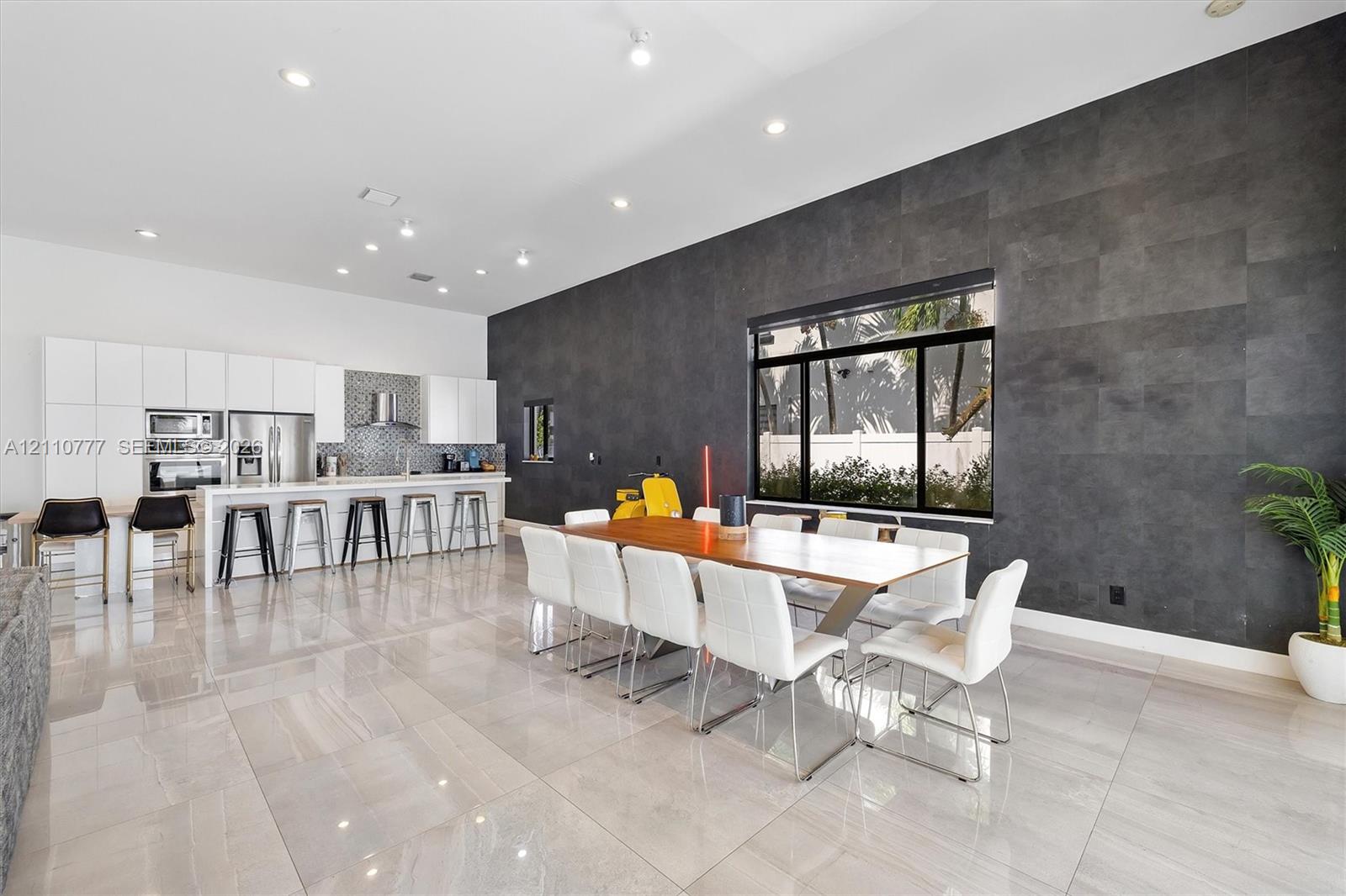 Modern kitchen and dining area featuring high-gloss white cabinetry, expansive stone tile flooring, a dark charcoal textured accent wall, recessed lighting, and built-in stainless steel wall ovens Modern kitchen and dining area featuring high-gloss white cabinetry, expansive stone tile flooring, a dark charcoal textured accent wall, recessed lighting, and built-in stainless steel wall ovens