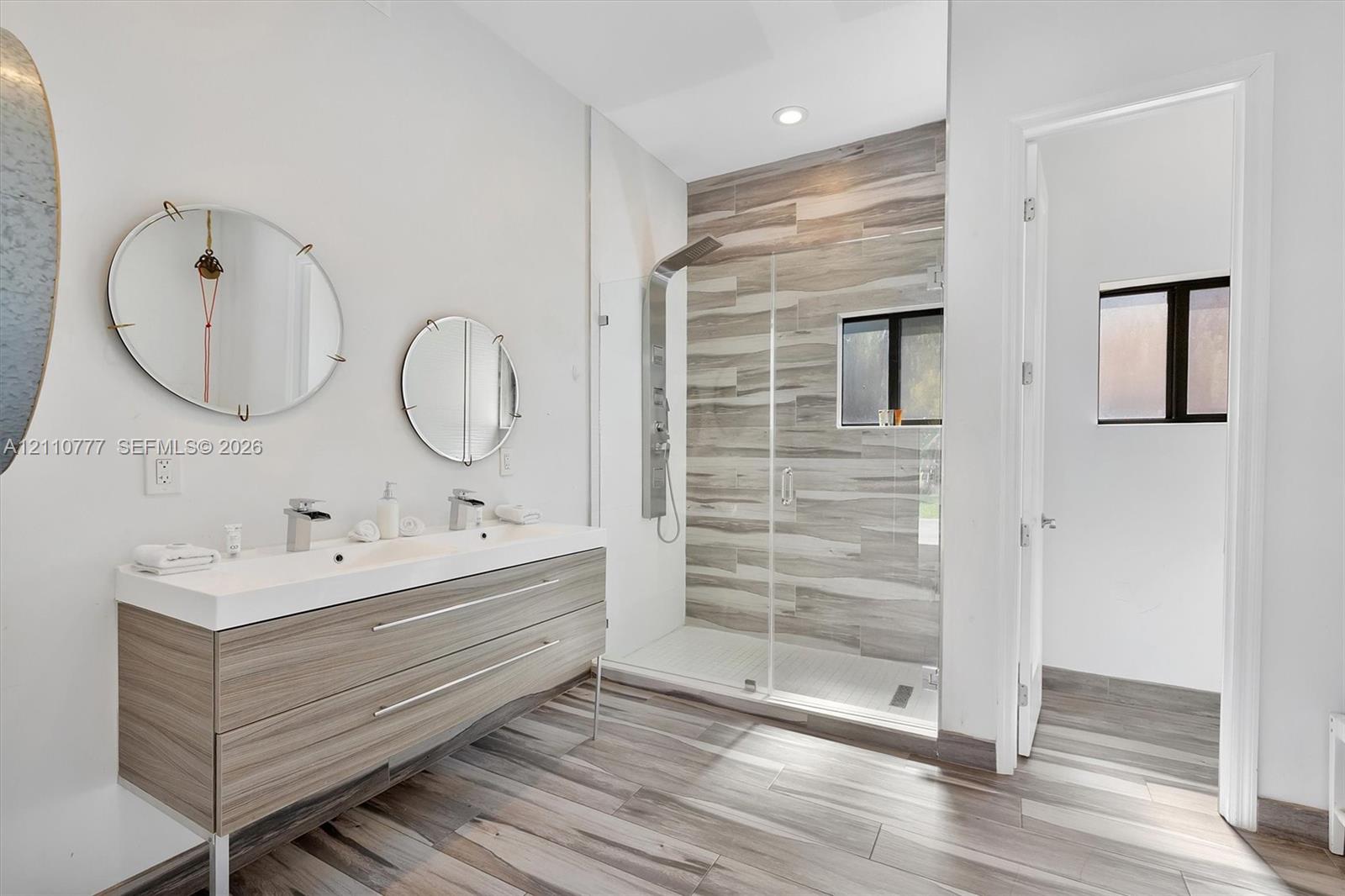Double vanity with integrated sinks, wall-mounted faucets, wood-finish cabinetry, walk-in shower with horizontal tile surround, and wood-finish flooring