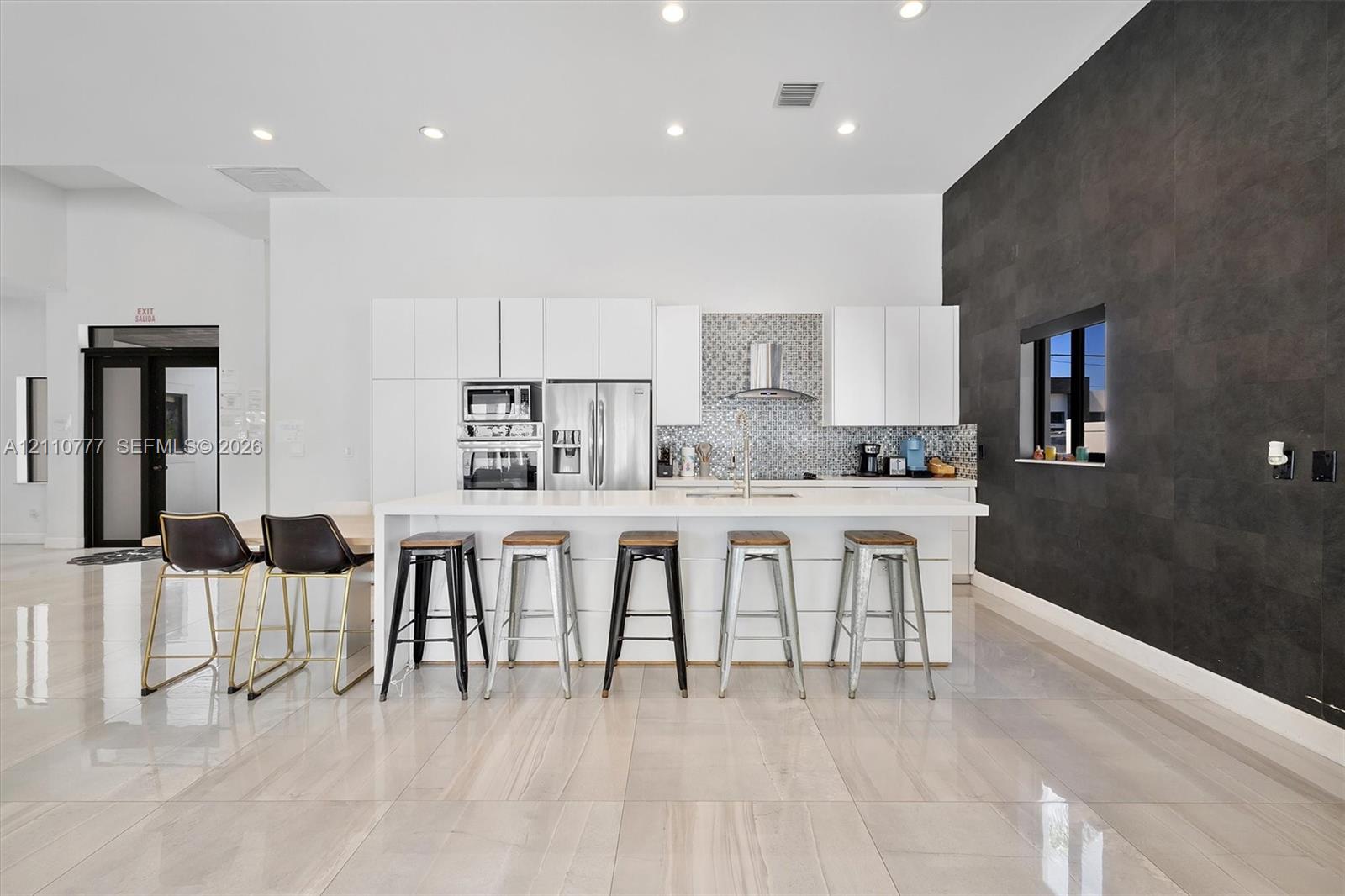 Modern kitchen featuring flat-panel white cabinetry, a waterfall-edge kitchen island, stone-finish tile flooring, a mosaic tile backsplash, and a dark tile accent wall Modern kitchen featuring flat-panel white cabinetry, a waterfall-edge kitchen island, stone-finish tile flooring, a mosaic tile backsplash, and a dark tile accent wall