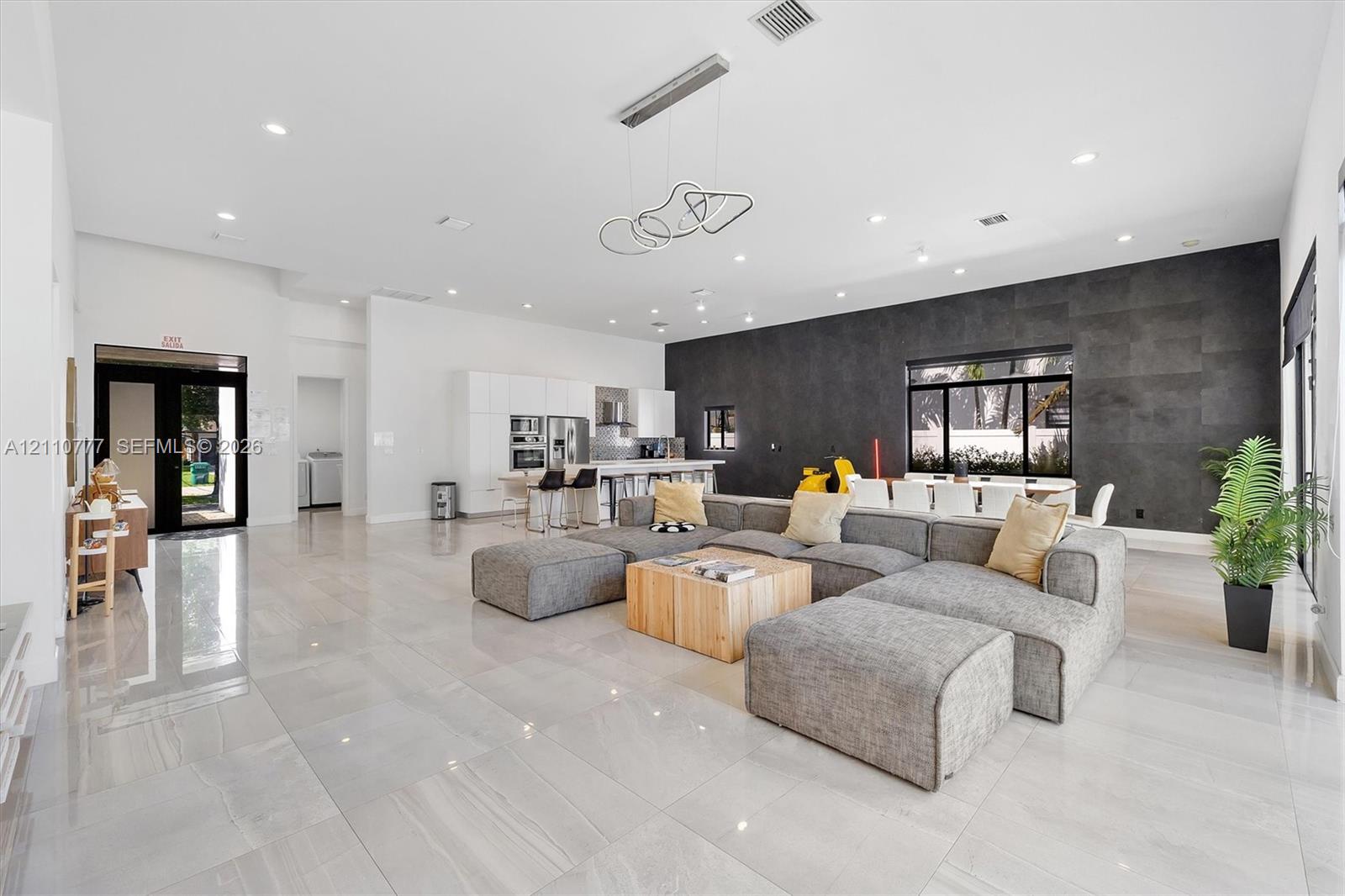 Open-concept living space featuring high ceilings with recessed lighting, large-format tile flooring, a charcoal stone-finish accent wall, and contemporary geometric pendant lighting Open-concept living space featuring high ceilings with recessed lighting, large-format tile flooring, a charcoal stone-finish accent wall, and contemporary geometric pendant lighting