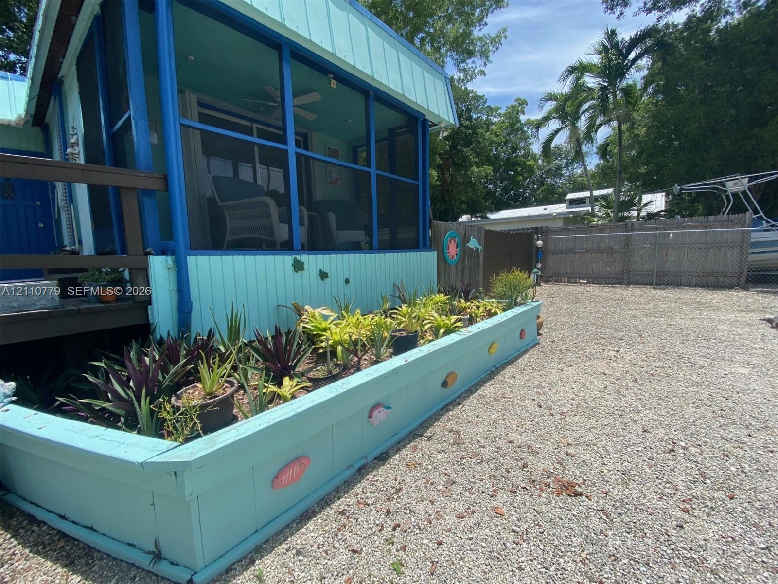 Screened porch with blue framing, elevated wooden deck, and a custom raised landscape planter box filled with assorted greenery, set against a gravel exterior surface