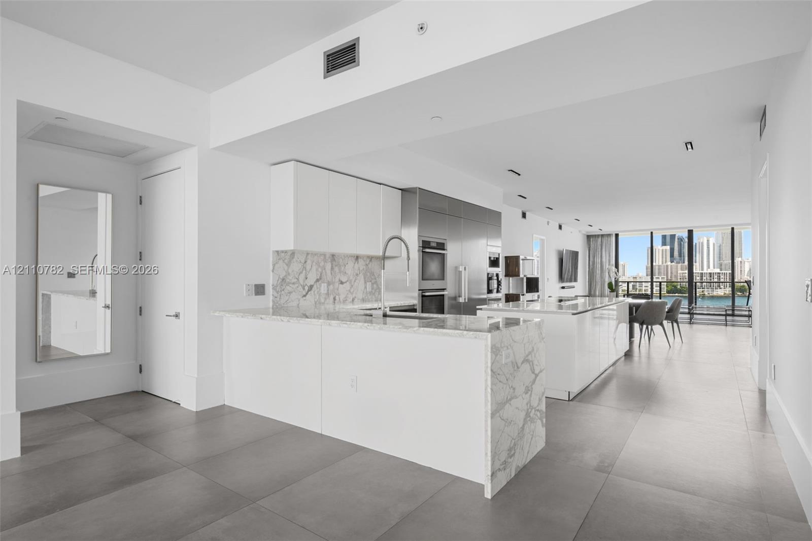 Kitchen featuring a stone-finish island with waterfall edge, white flat-panel cabinetry, stainless steel built-in appliances, and large-format floor tiling