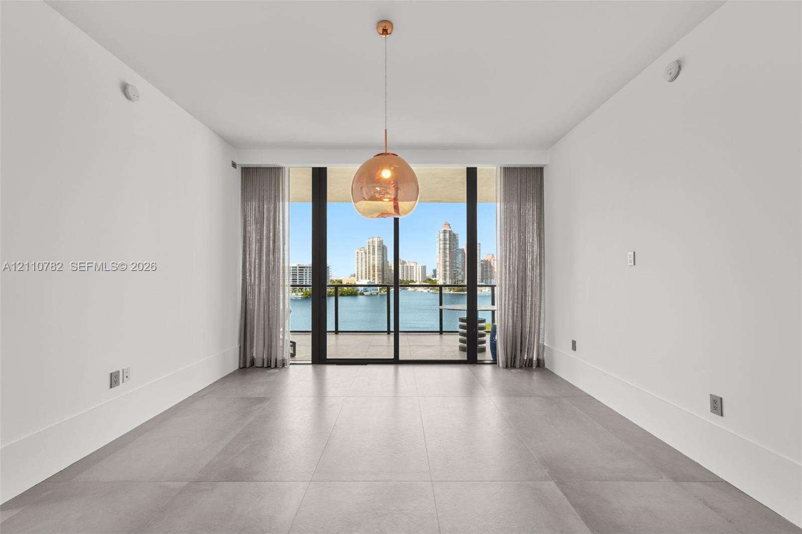 Large-format tile flooring, oversized sliding glass door, spherical glass pendant light, and balcony with water and city skyline backdrop