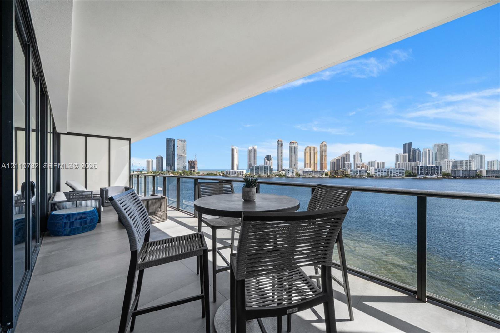 Expansive covered balcony featuring large-format stone-finish flooring, full-height glass doors, and a sleek glass railing with a metal frame