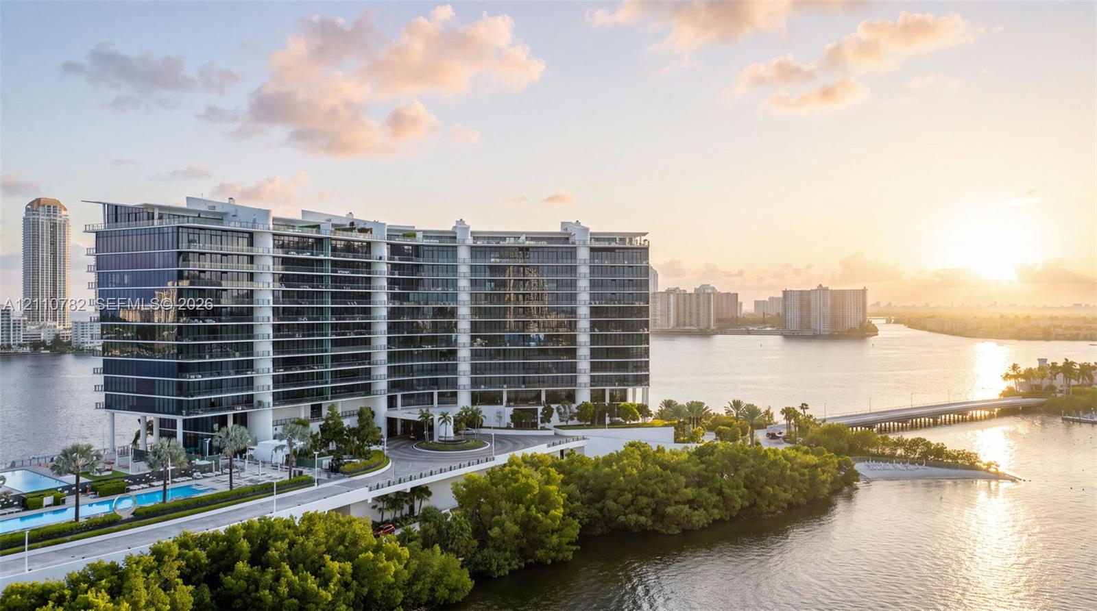 High-rise residential building featuring floor-to-ceiling glass paneling, wrap-around balconies, a porte-cochre entryway, and an outdoor swimming pool, situated on a waterfront lot with dense perimeter foliage