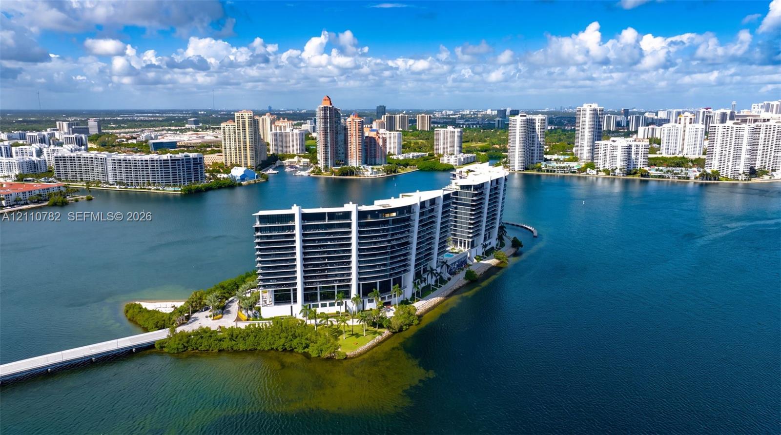 Waterfront high-rise building situated on a private peninsula, featuring wraparound balconies, an outdoor swimming pool, lush landscaping, and direct access via a private bridge Waterfront high-rise building situated on a private peninsula, featuring wraparound balconies, an outdoor swimming pool, lush landscaping, and direct access via a private bridge