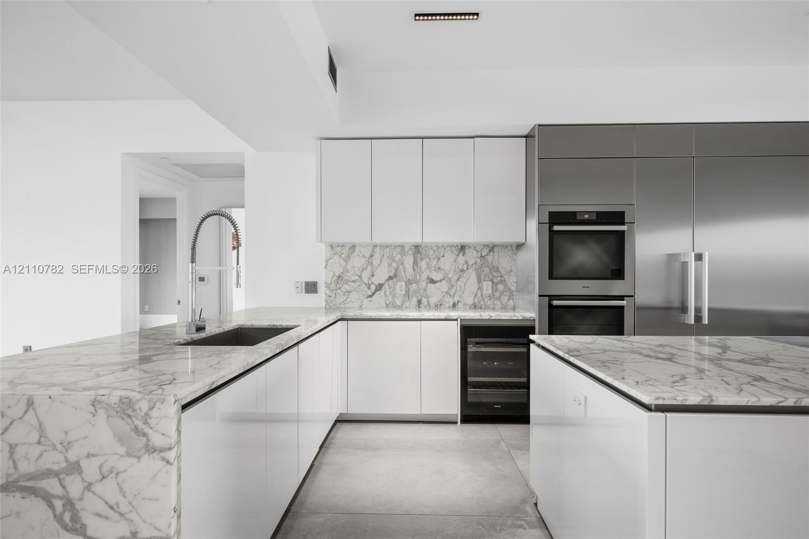 Kitchen featuring white flat-panel cabinetry, stone countertops with waterfall edge, full-height stone backsplash, stainless steel double wall ovens, and an under-counter beverage cooler